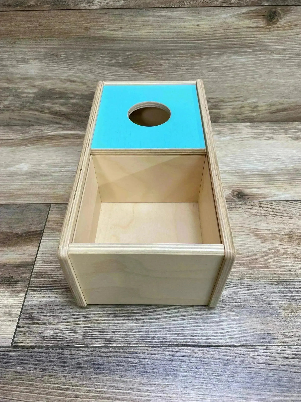 Top-down angled view of Lovevery sliding-top box showing turquoise lid with round hole and open compartment on wood floor