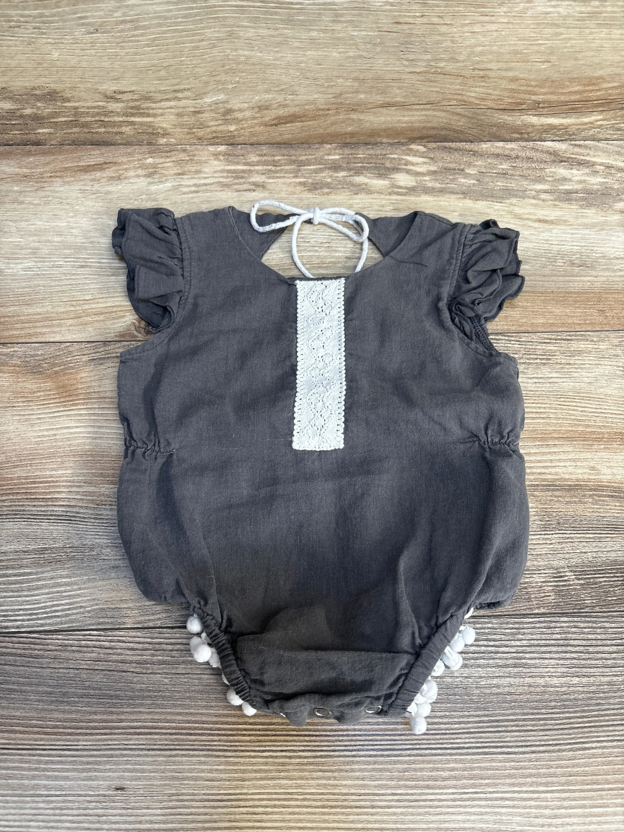 Gray baby romper with ruffled sleeves, white lace trim, and pom-pom hem on wood floor