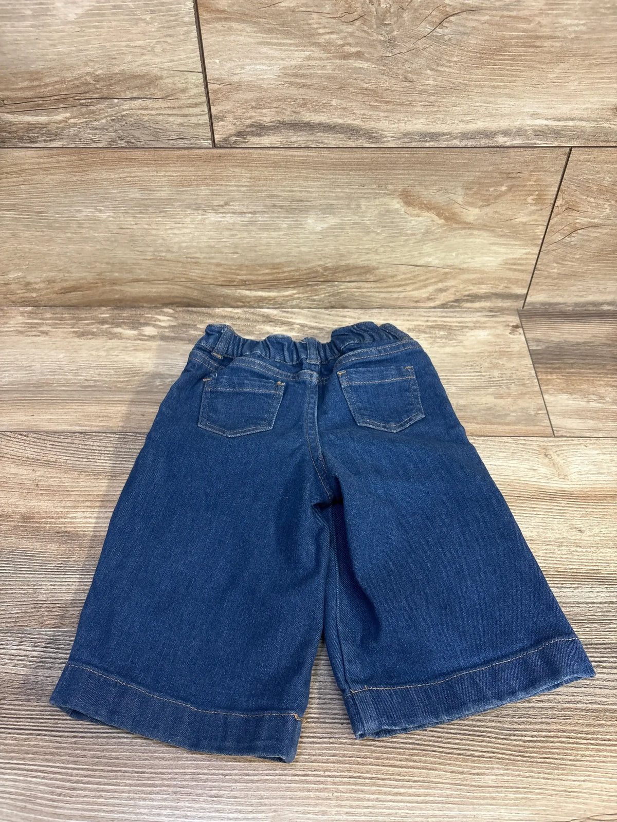 Flat lay of toddler wide-leg denim jeans showing pleated front, elastic waistband and back patch pockets in medium wash on wood floor