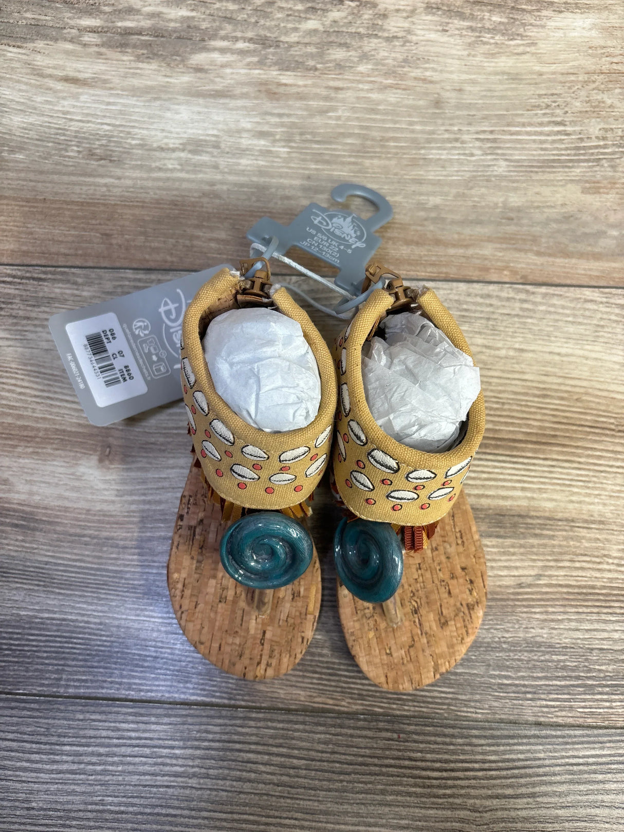 Top view of toddler Moana sandals with cork soles, yellow ankle straps and teal spiral toe decorations on wooden surface