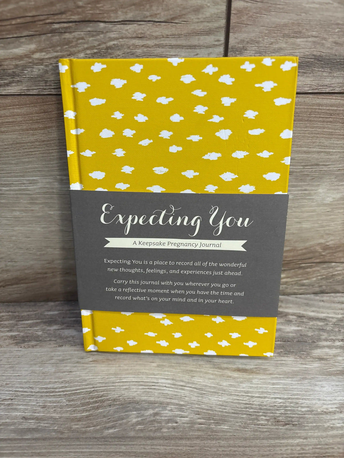 Front view of Expecting You pregnancy journal with yellow hardcover, white cloud pattern, and gray belly band on wood backdrop