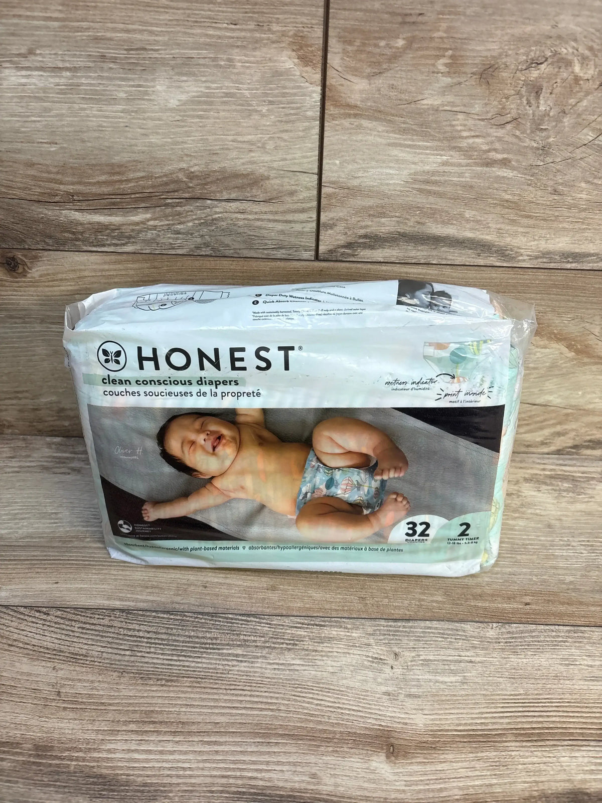 Front view of Honest Clean Conscious diapers pack (size 2, 32 count) showing baby photo and Tummy Timer badge on wood floor