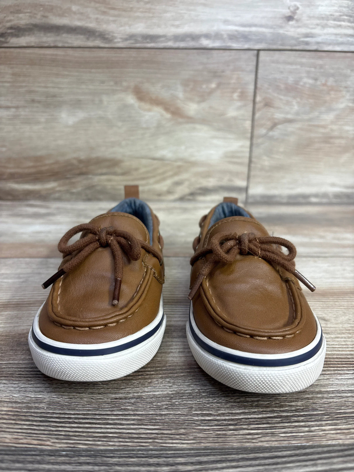 Pair of brown toddler boat shoes with white rubber soles and brown laces on wooden floor