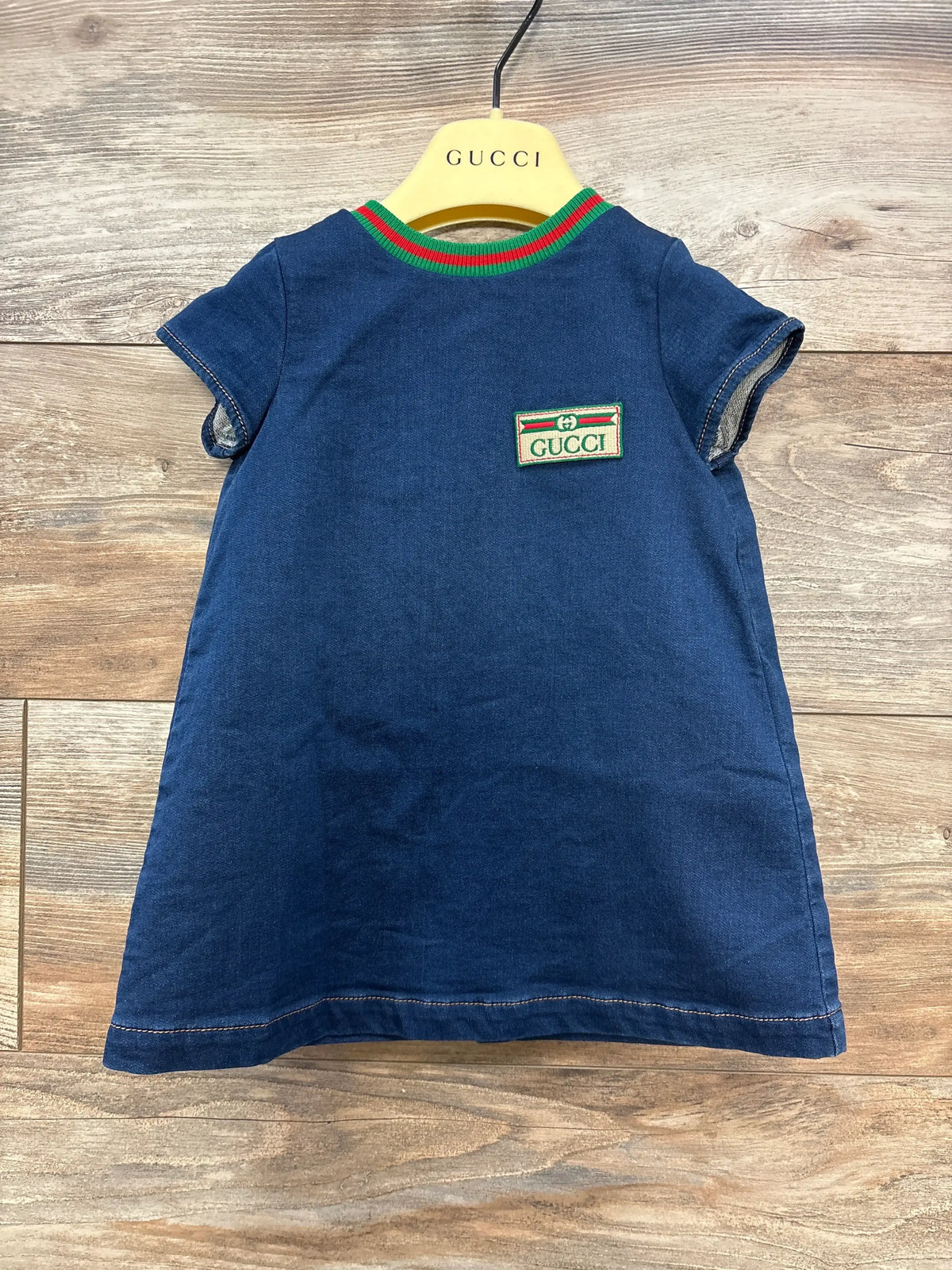 Front view of Gucci denim dress in blue with green and red striped collar and Gucci patch logo on yellow Gucci hanger