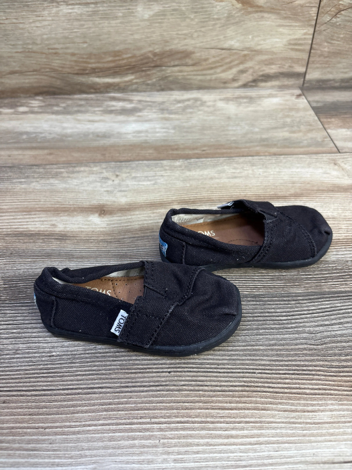 Angled top view of Toms Tiny Alpargata toddler shoes, black canvas slip-on with TOMS tag and rubber soles on wood background