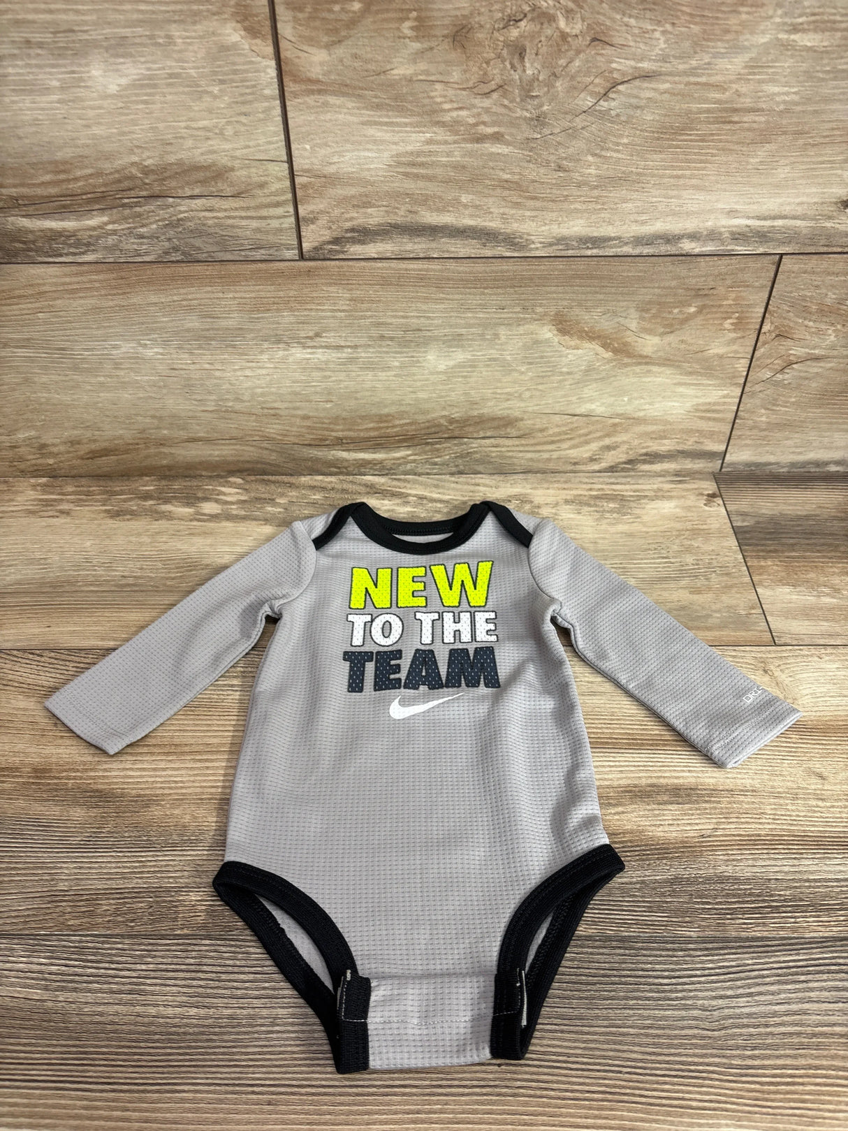 Flat lay of grey Nike Dri-Fit long-sleeve bodysuit showing NEW TO THE TEAM text, black trim, and snaps on wood background