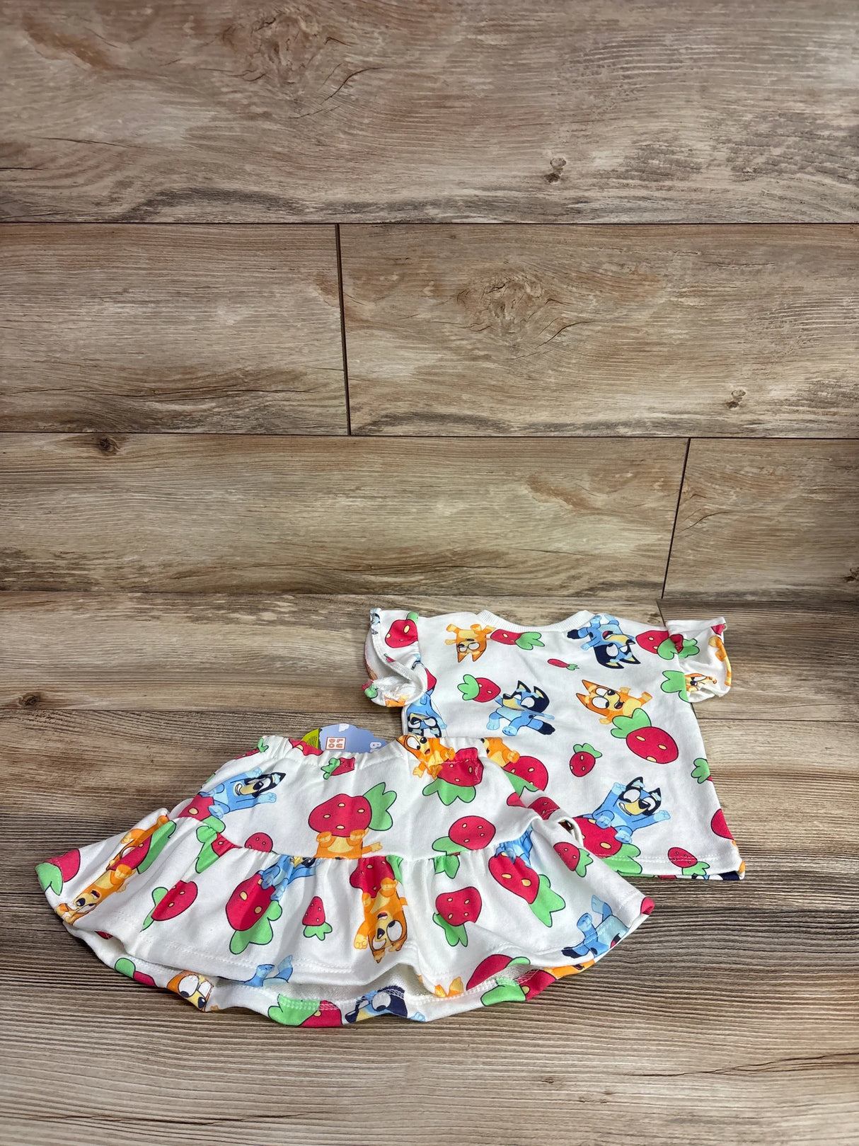 Flat lay front view of white toddler two-piece top and ruffled skort set showcasing Bluey characters and red strawberry print on wood background