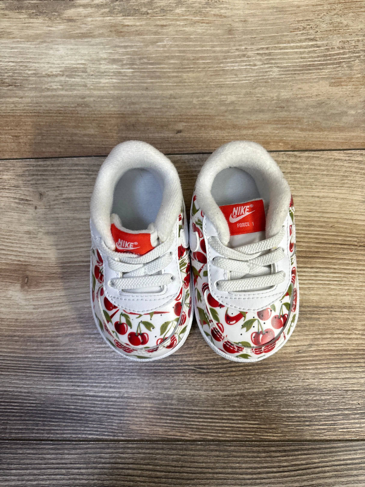 Top view of Nike Air Force 1 crib shoes showcasing red cherry print and red Nike tongue label on wood floor background
