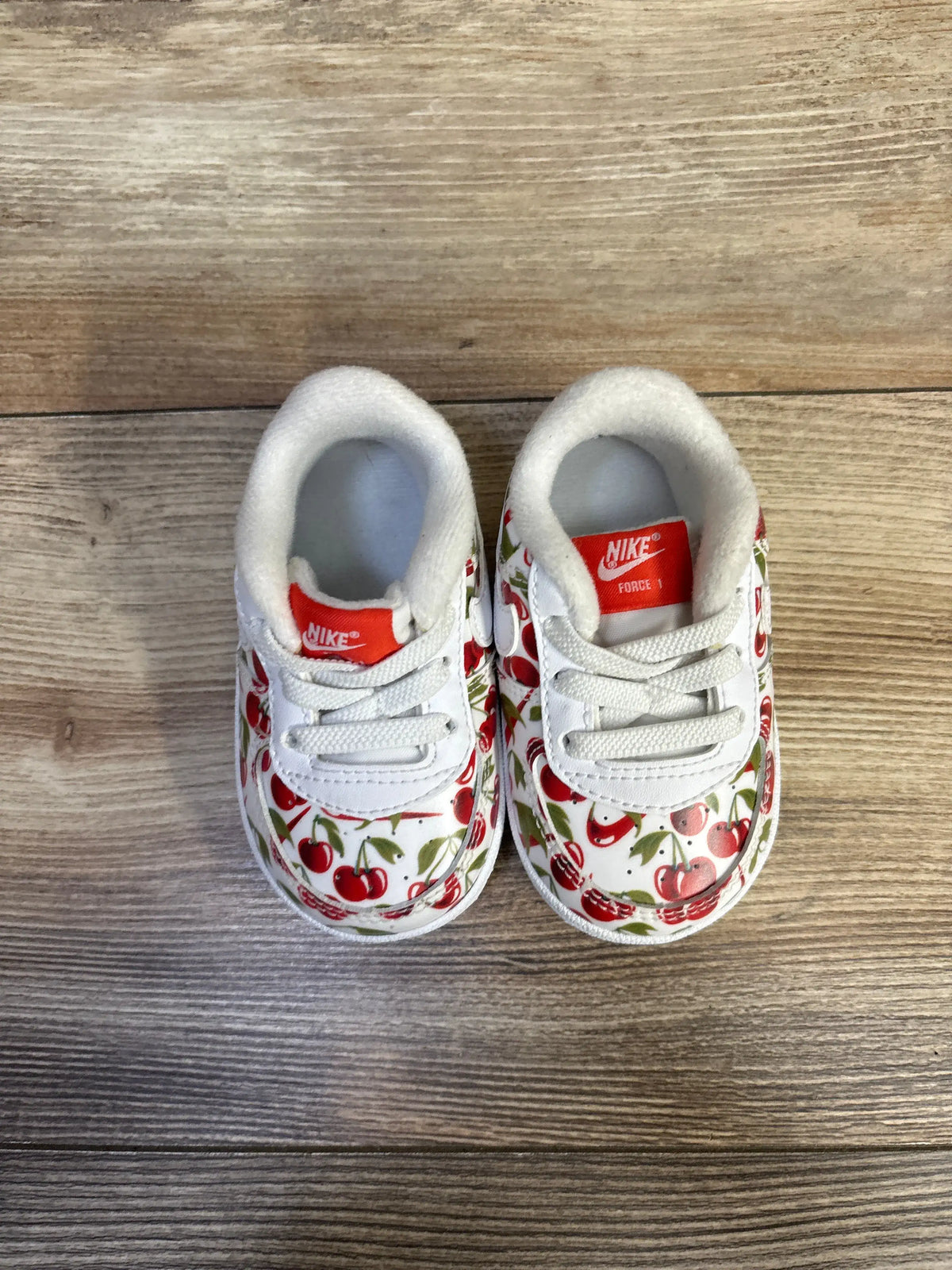 Top view of Nike Air Force 1 crib shoes showcasing red cherry print and red Nike tongue label on wood floor background