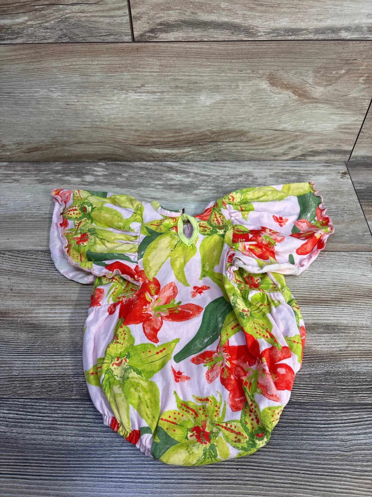 Top view of Burt’s Bees floral romper in pink with flutter sleeves and gathered leg openings on a wood-look floor