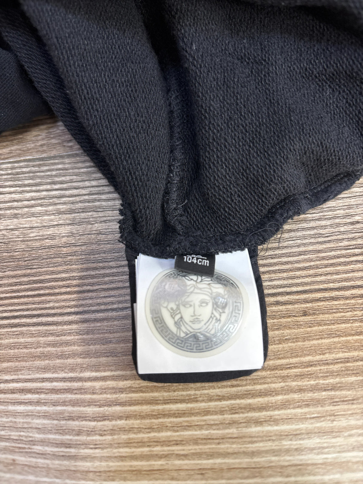 Black knit fabric with Versace Medusa logo clothing tag and size label 104cm on wooden surface