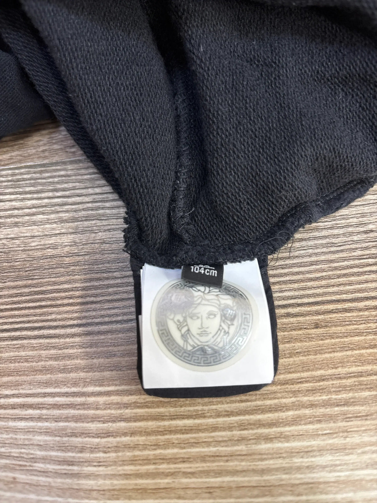 Black knit fabric with Versace Medusa logo clothing tag and size label 104cm on wooden surface