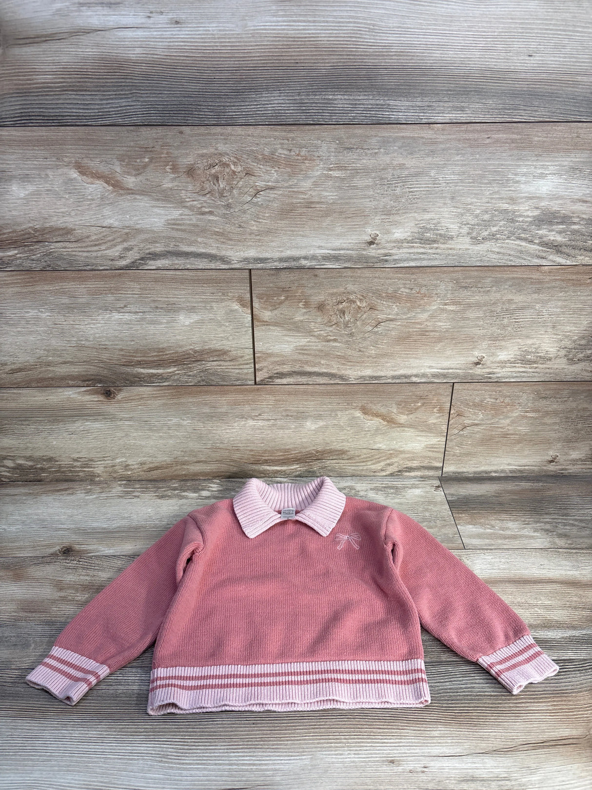 Front flat lay of pink Carters sweater 5T showcasing white ribbed collar, embroidered bow and striped hem on wood background