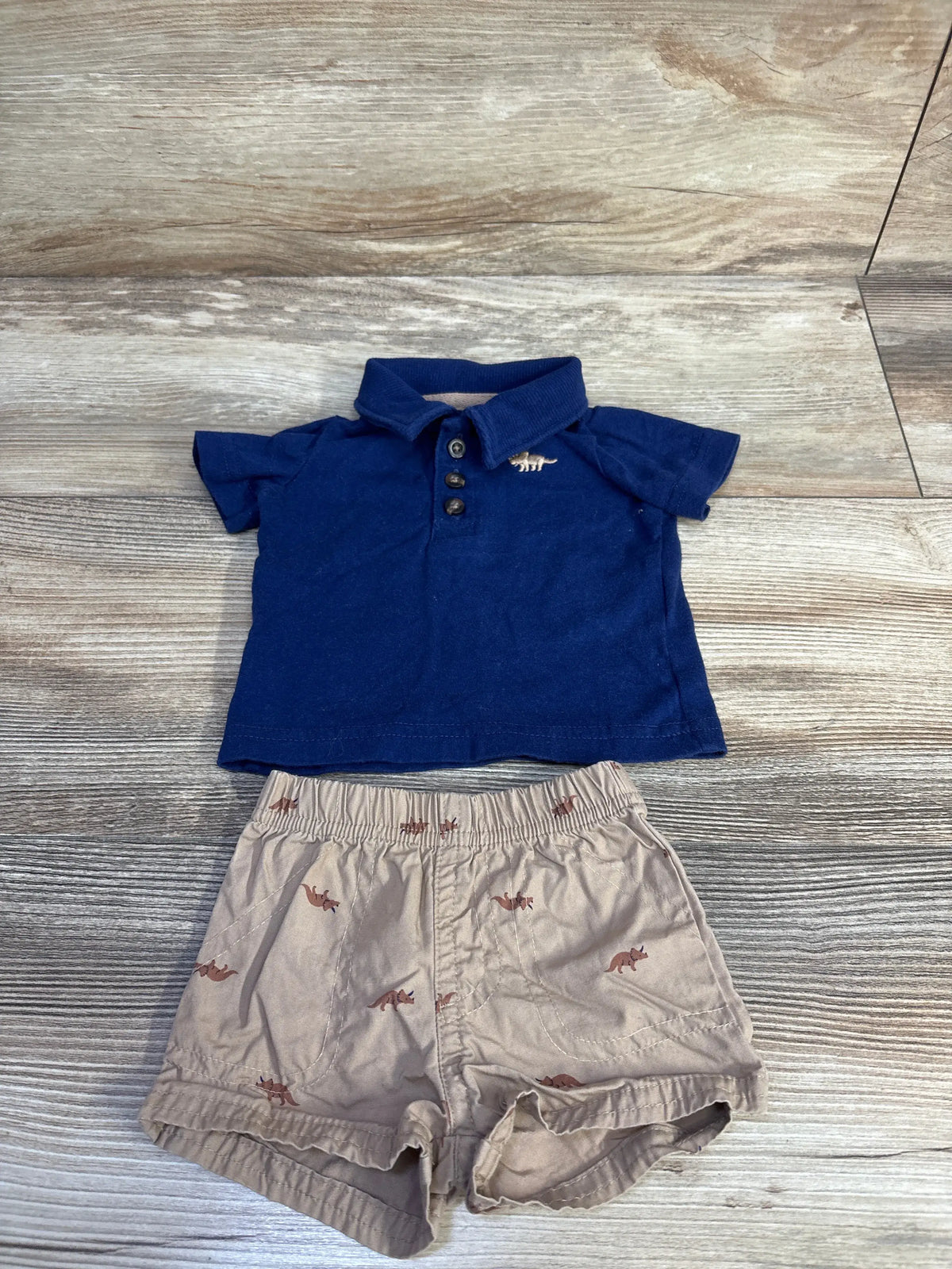 Top view of Carter’s two-piece newborn outfit showcasing a navy collared polo and dinosaur-print shorts on wood-look flooring
