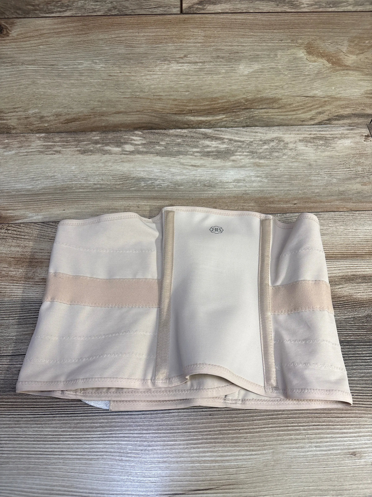 Front view of the Belly Bandit Luxe Postpartum Belly Wrap showcasing beige layered panels and stitched edges on a wood surface