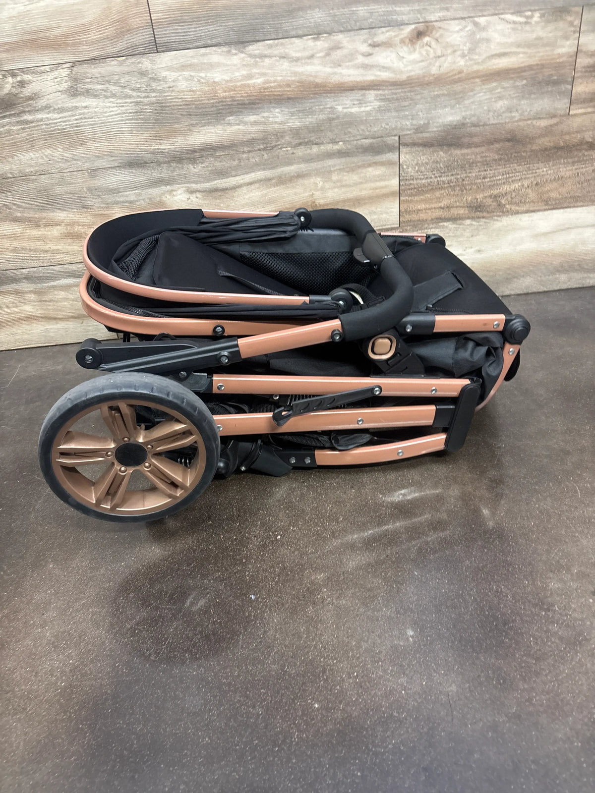 Side view of folded black 2-in-1 stroller pushchair showcasing rose gold frame and large wheel on a wood and concrete floor
