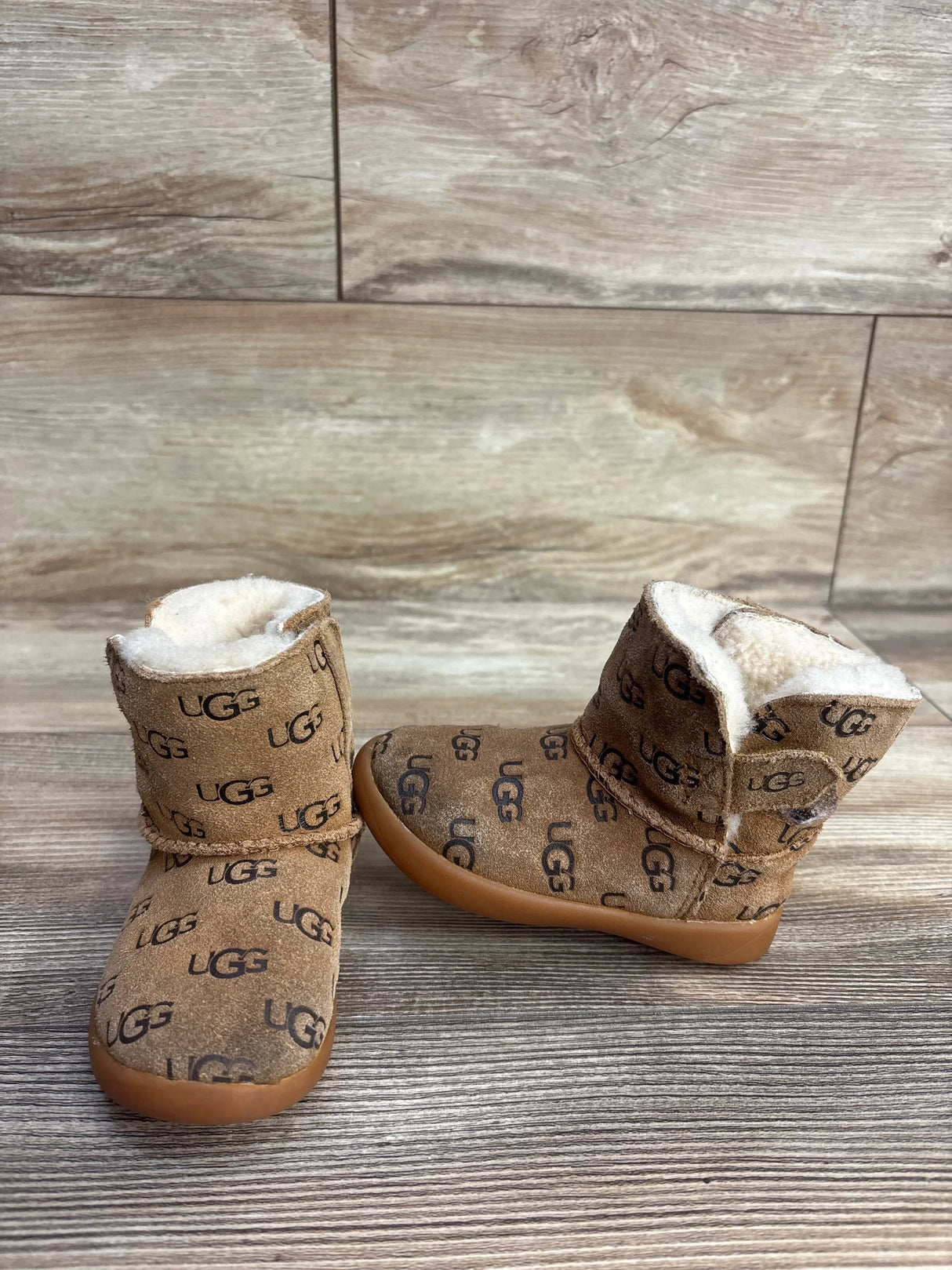 Pair of brown UGG baby boots with fleece lining and repeating logo pattern on wooden surface