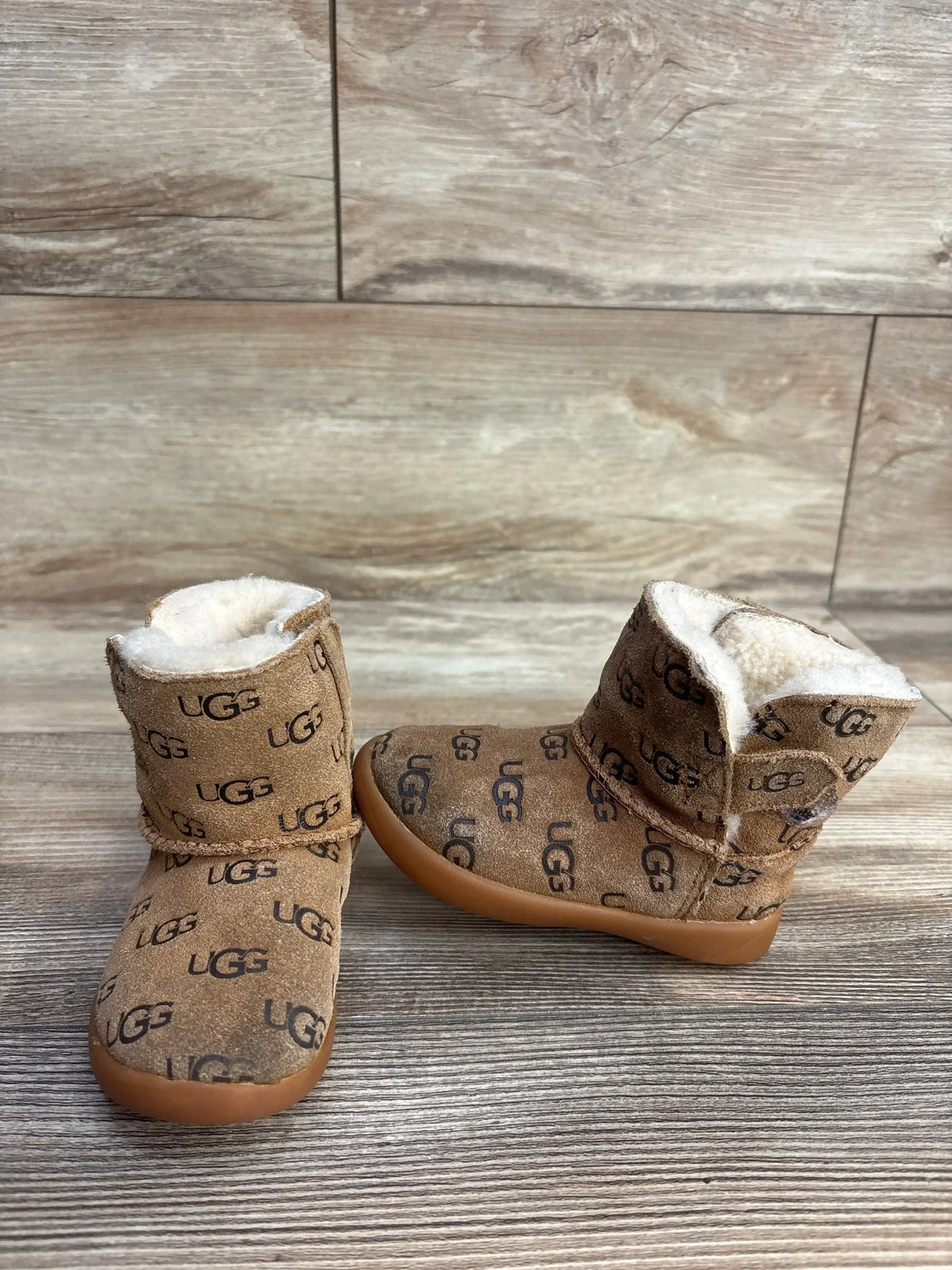 Pair of brown UGG baby boots with fleece lining and repeating logo pattern on wooden surface