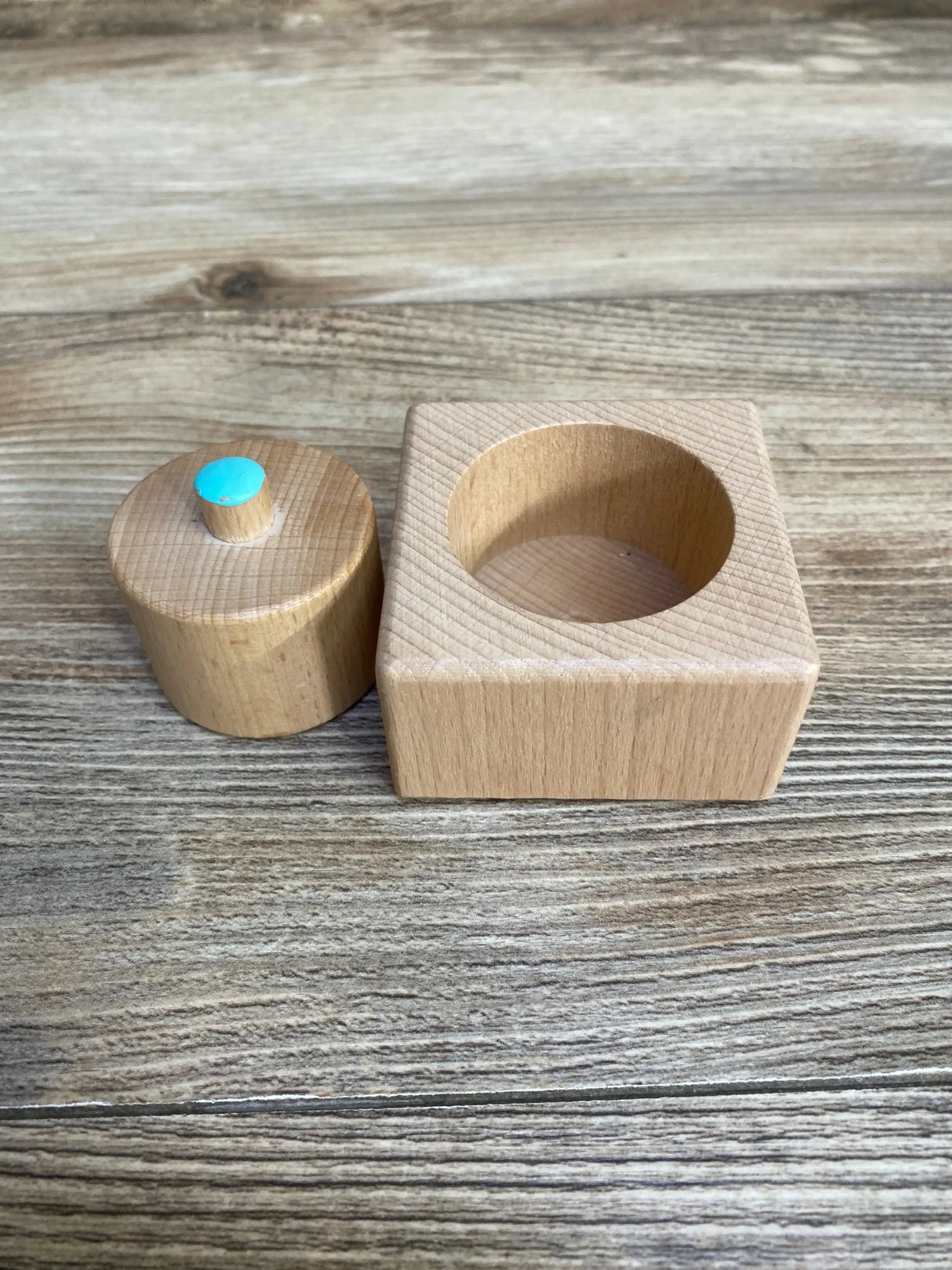 Front view of Pincer Puzzle showing wooden peg with turquoise top beside square base with hole on wood background