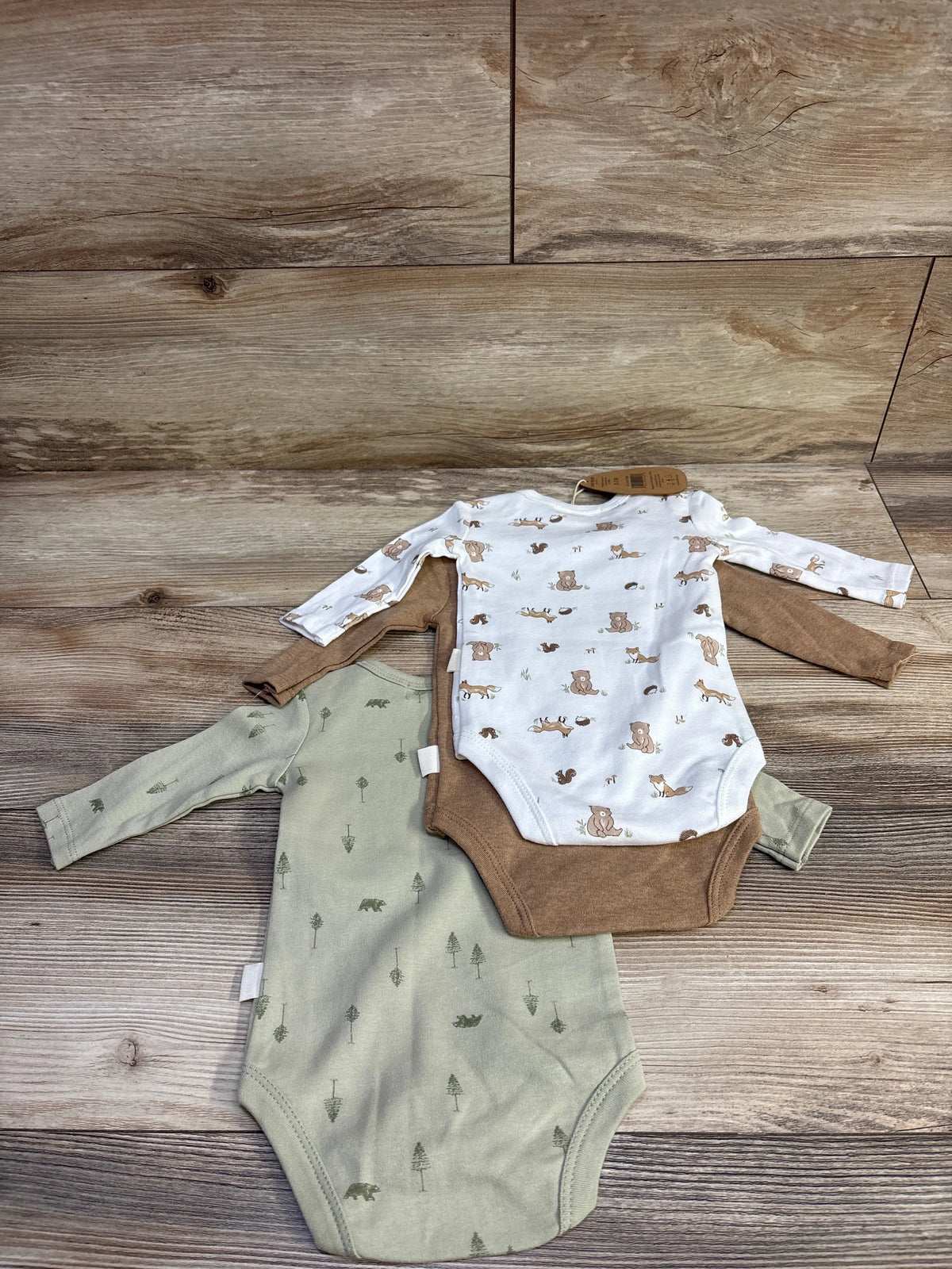 Flat lay of 3-pack organic long-sleeve baby bodysuits showing white animal-print, brown solid, and sage green tree-print on wood background