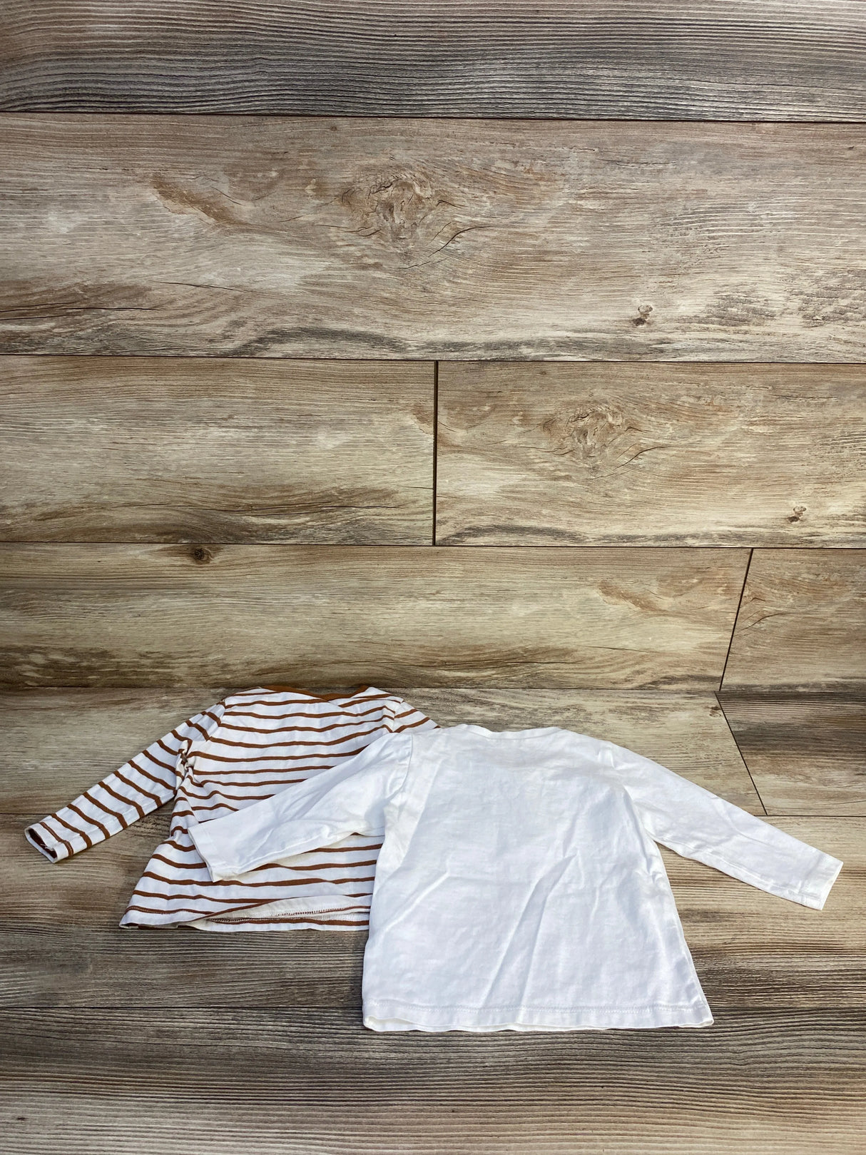 Two long sleeve toddler shirts, one white and one brown striped, on wood background