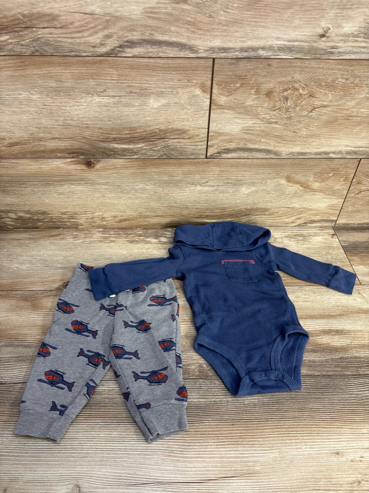 Baby clothes set with navy blue hooded bodysuit and grey pants featuring red and blue helicopter prints on wood background