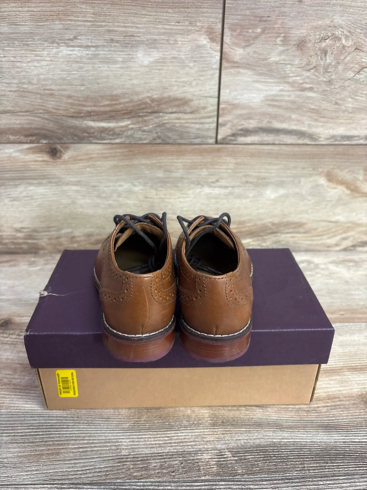 Rear of Johnston & Murphy LK Conard brown cap-toe shoes with brogue detailing and black laces on shoebox and wood background