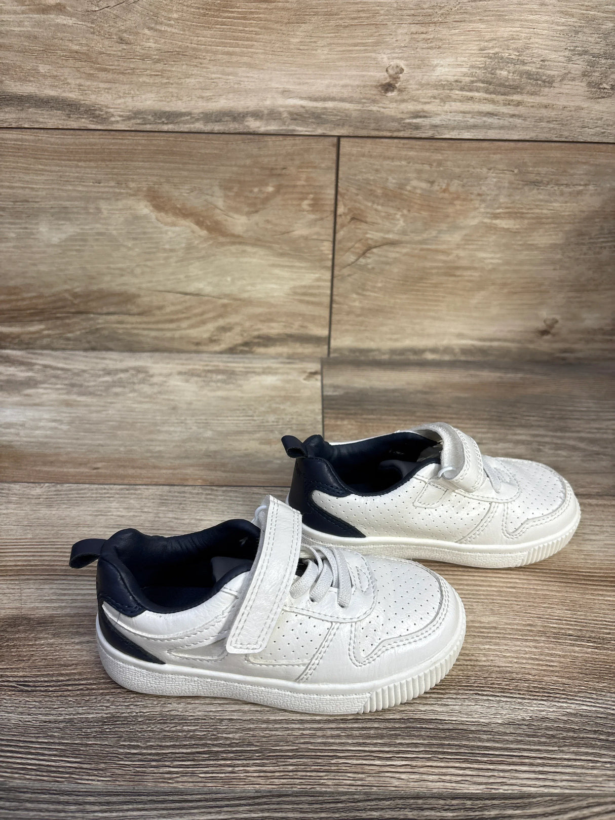 Side view of Carter's Every Step Port sneakers showing white perforated upper, navy heel, and Velcro strap on wood backdrop