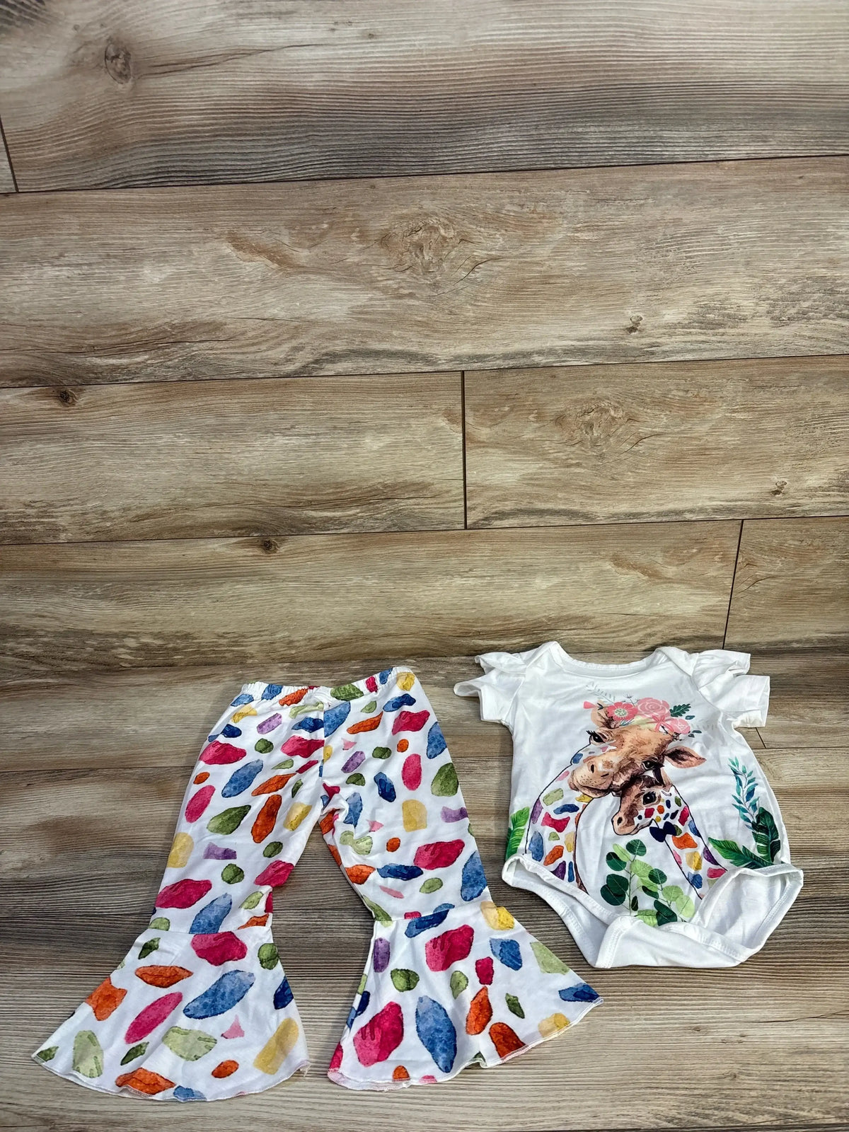 Flat lay of white giraffe-print bodysuit and matching colorful spotted bell-bottom pants on light wood floor