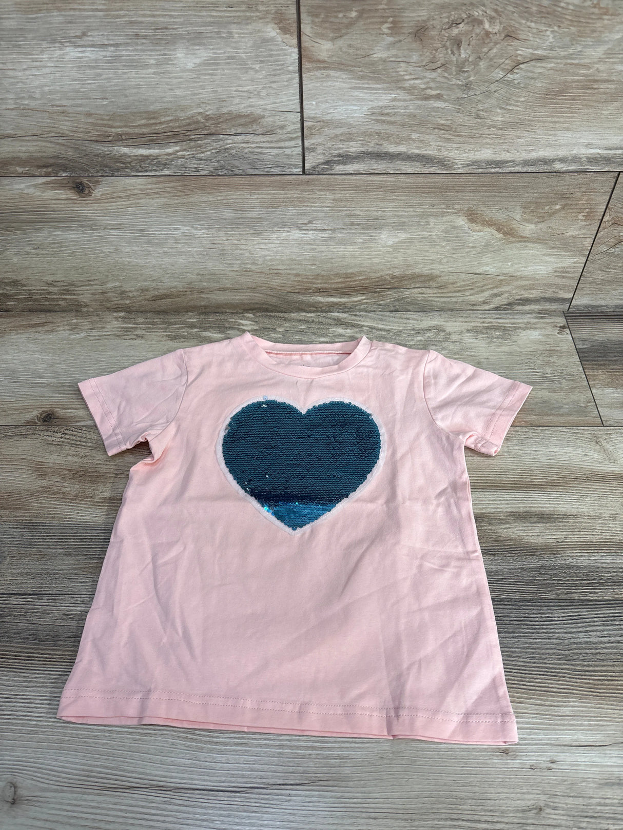 Light pink children’s short-sleeve t-shirt with large blue sequin heart on front, on wood floor