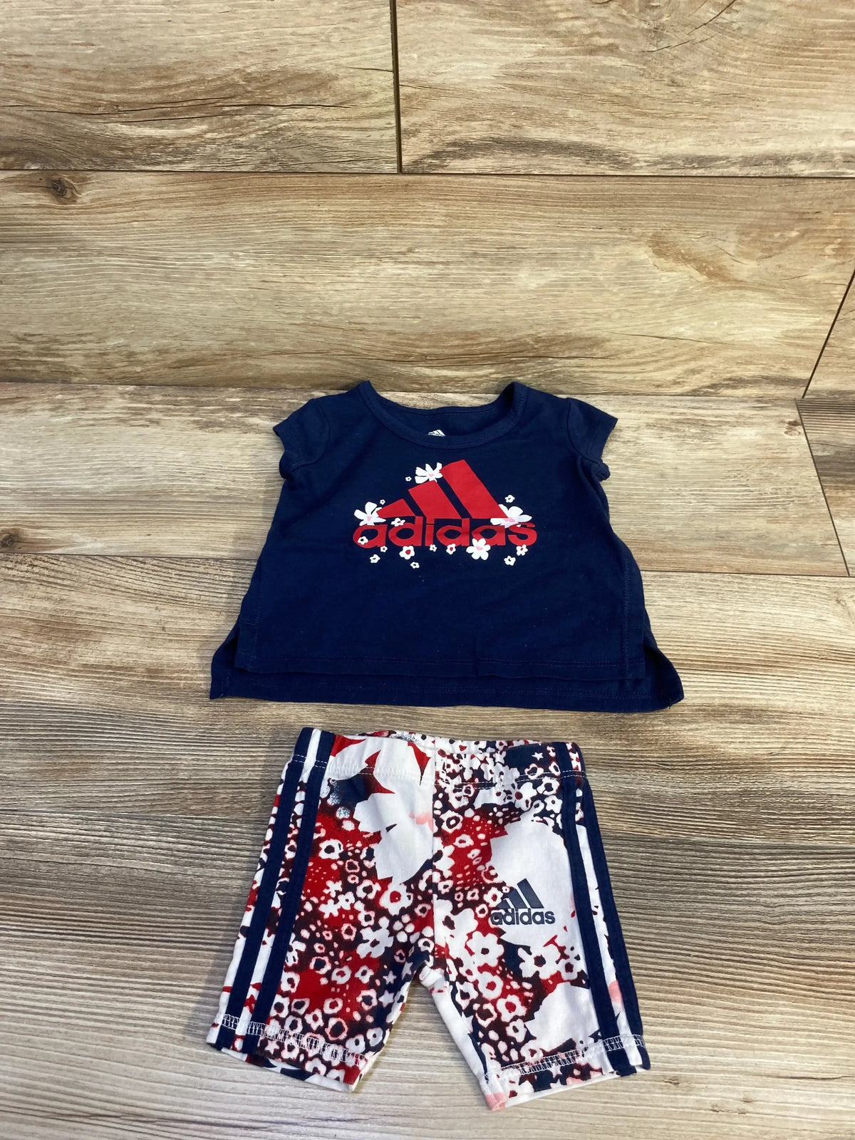 Flat-lay of navy baby Adidas 2-piece: tee with red floral Adidas logo and red-white floral shorts with navy stripes on wood