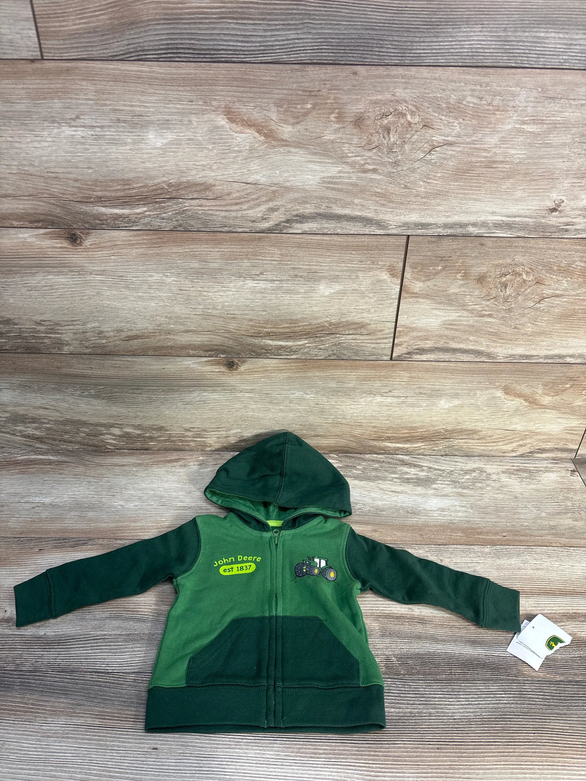 Front view of a green John Deere full-zip hoodie showcasing a hood, tractor graphic, and front pockets on a wood floor