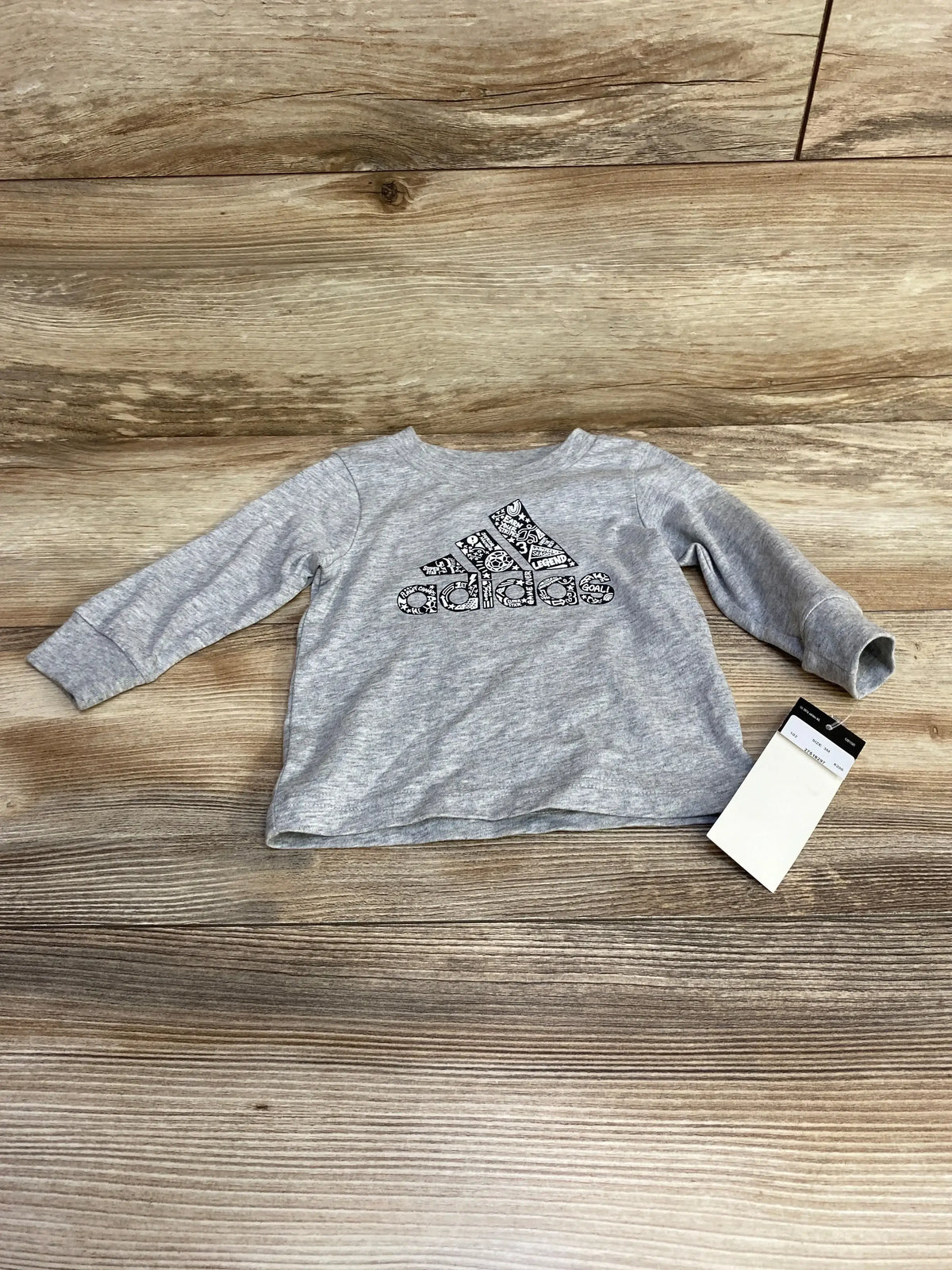 Flat lay front view of grey Adidas long-sleeve baby shirt (3M) showing patterned black Adidas logo and attached tag on wood background