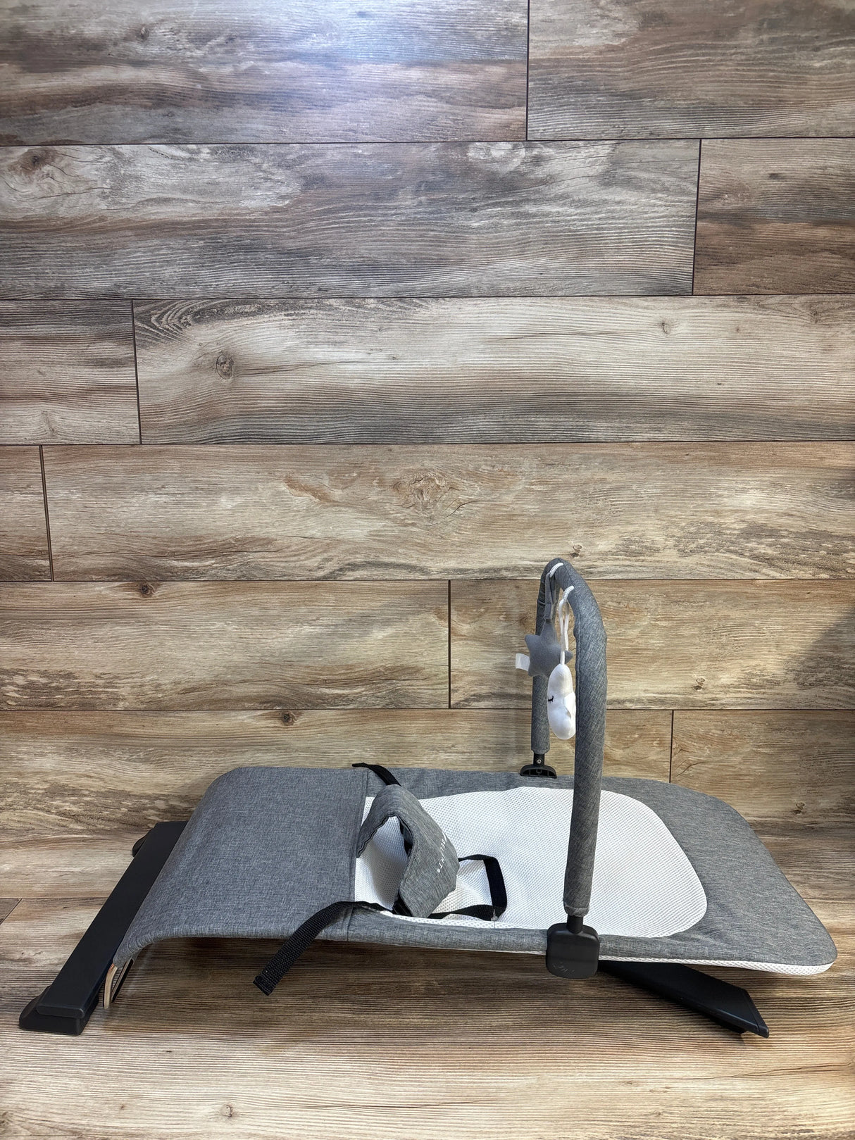 Side view of used gray baby bouncer seat showcasing reclined frame harness straps and folded toy bar on wood plank floor