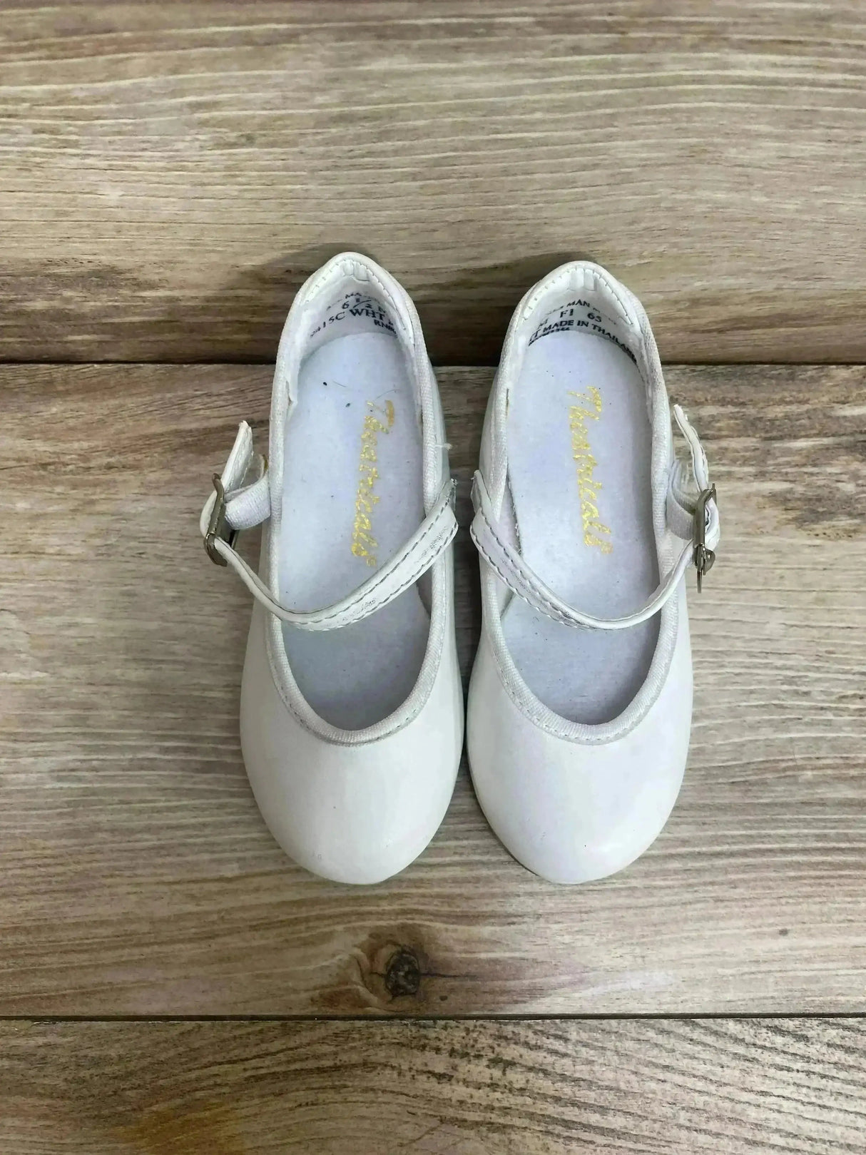Top-down view of white toddler tap buckle shoes showcasing Mary Jane strap and metal buckle on wooden floor background