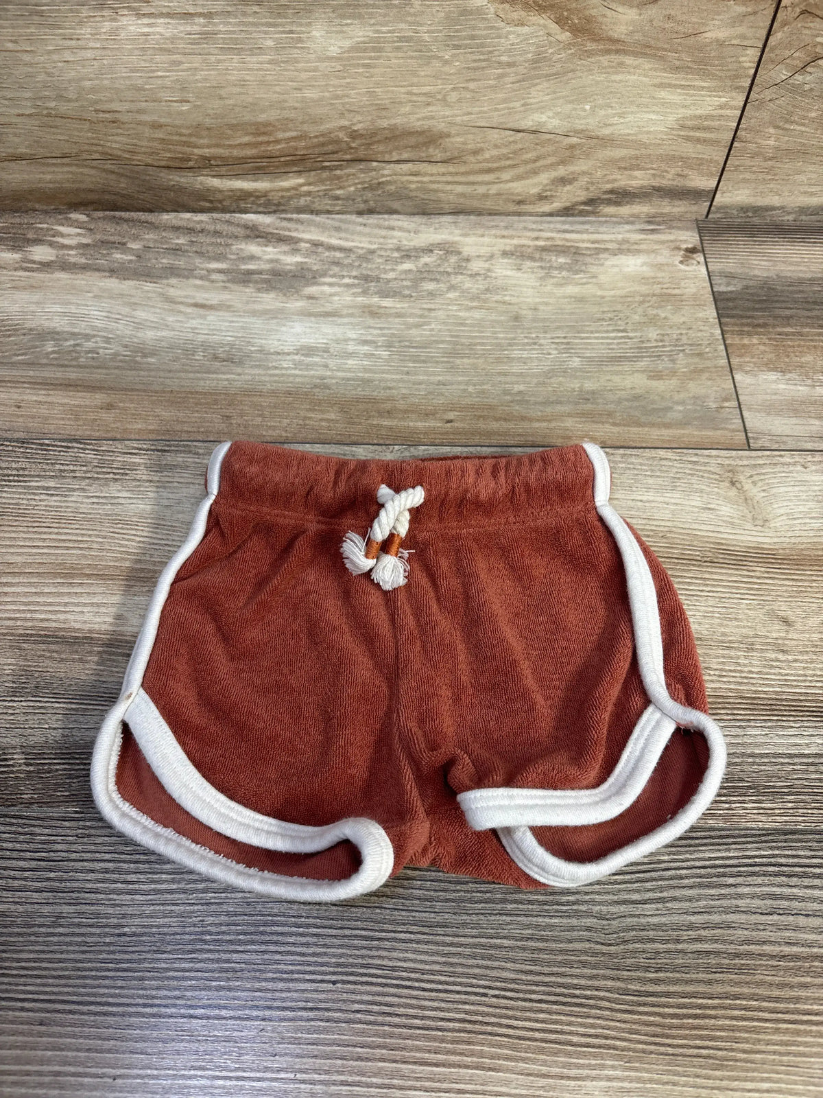 Front view of Little Co. terry dolphin shorts in Adobe Cup, size 6m, with white trim and drawstring elastic waistband on wood