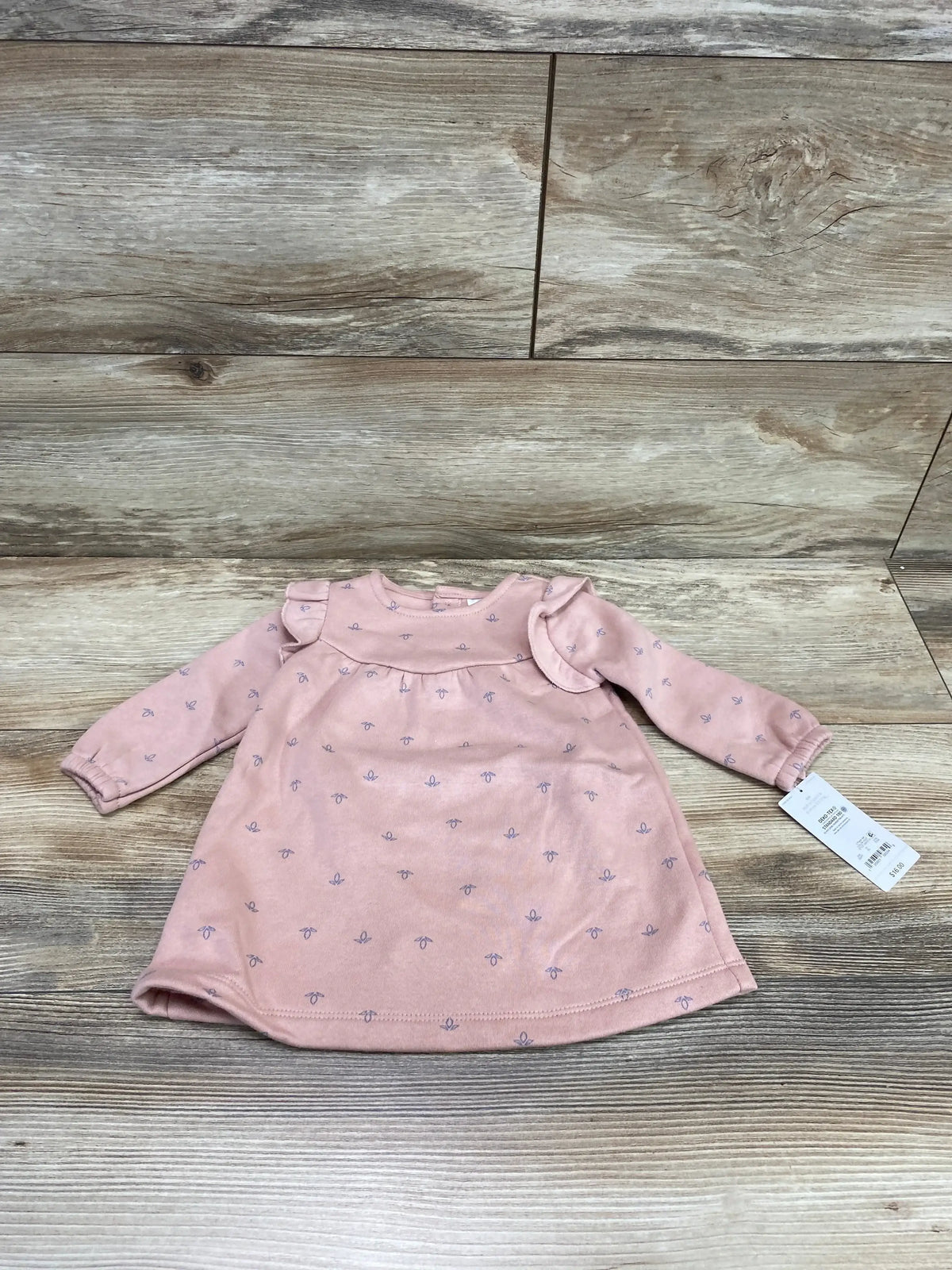 Flat lay of pink baby sweatshirt dress showcasing ruffle shoulder and tiny lavender floral print on wood background