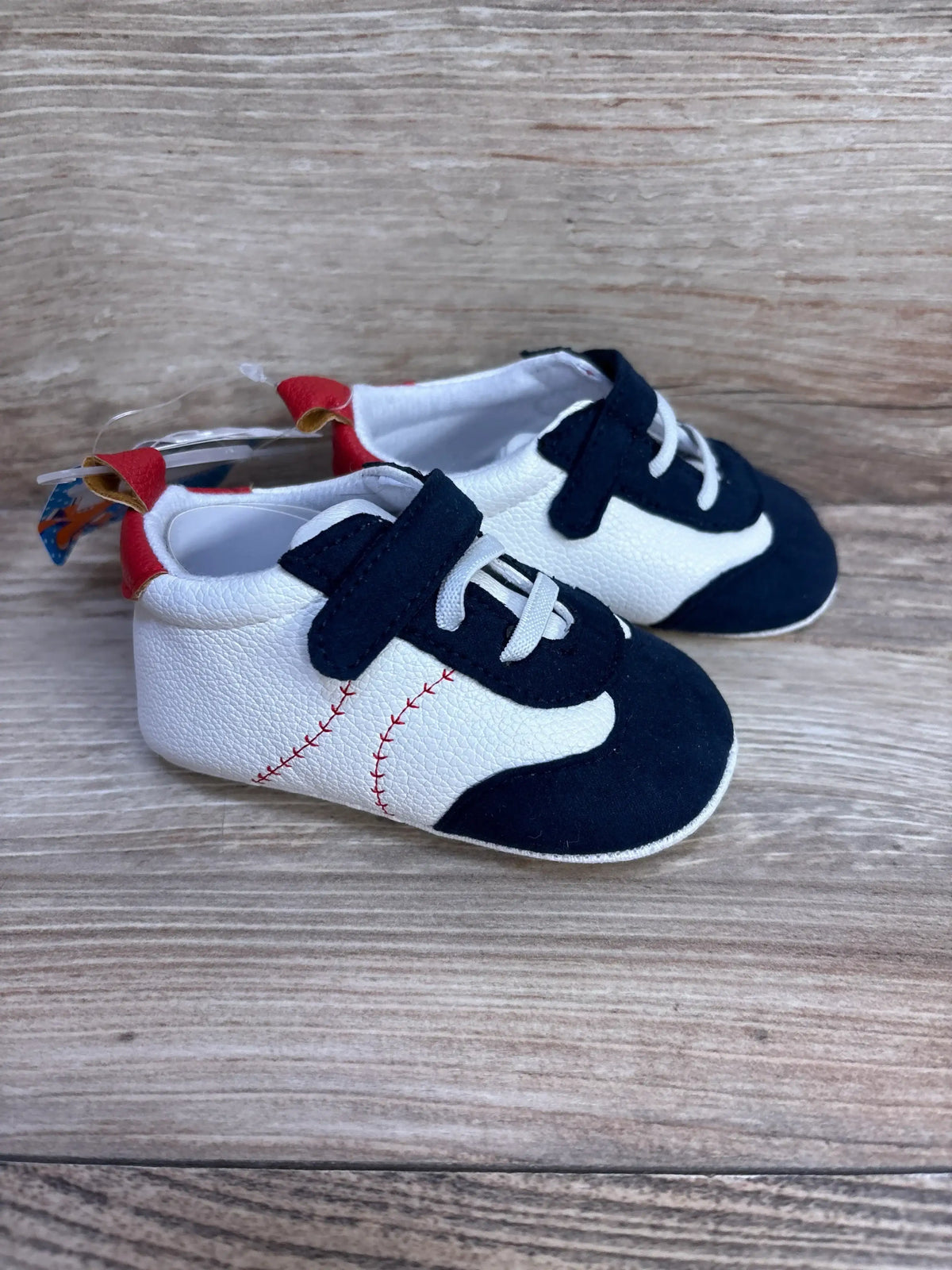 Angled side view of white and navy baby baseball shoes showing red baseball-stitch detail and Velcro strap on wood background