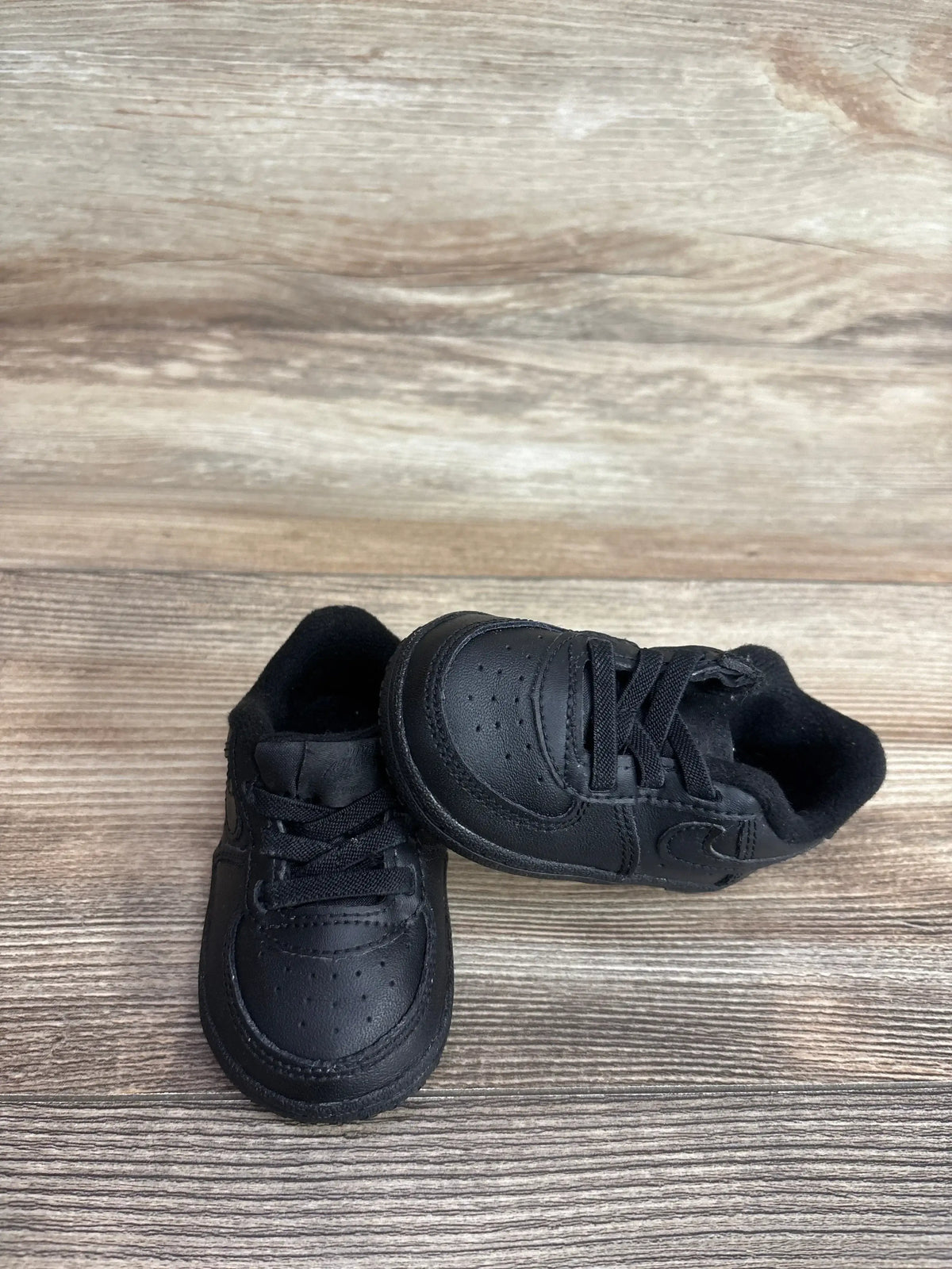 Angled view of Nike Air Force 1 Crib Shoes in black showcasing perforated leather upper and swoosh logo on wood plank floor