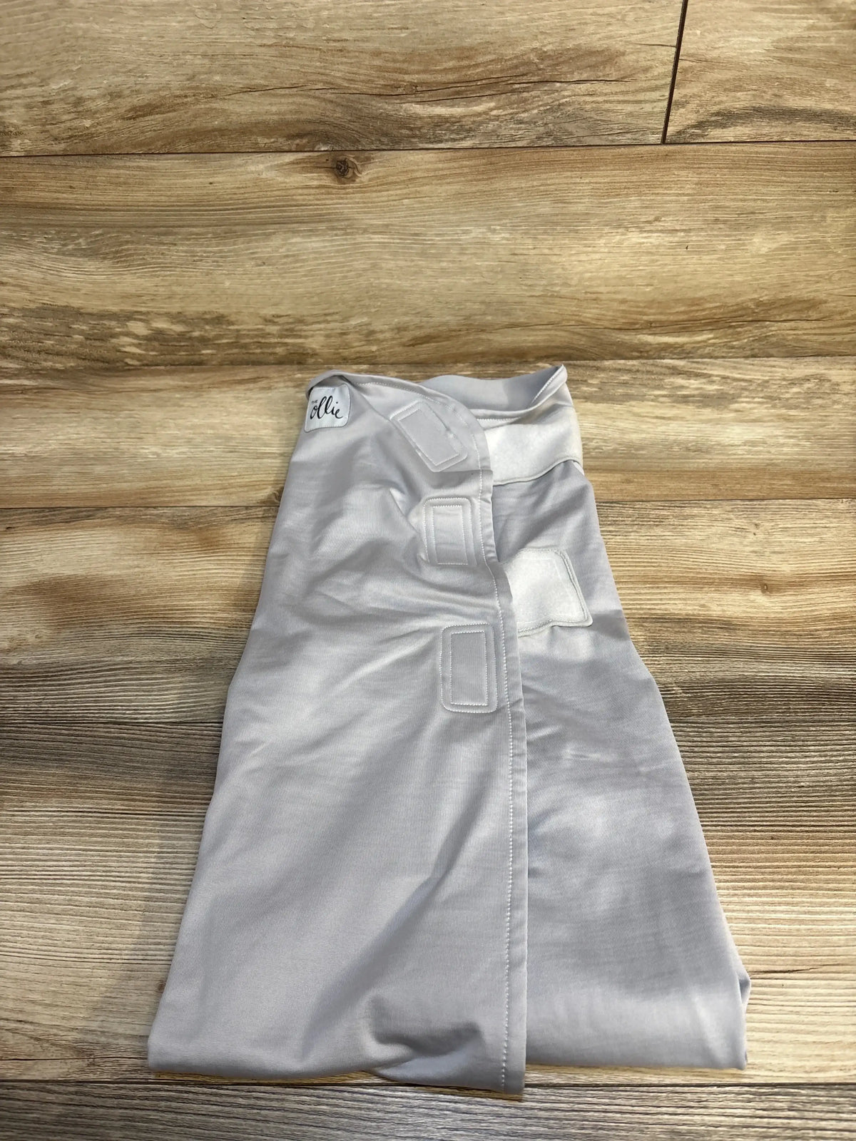Top-down view of The Ollie World swaddle in grey showcasing three Velcro closures on a wooden floor