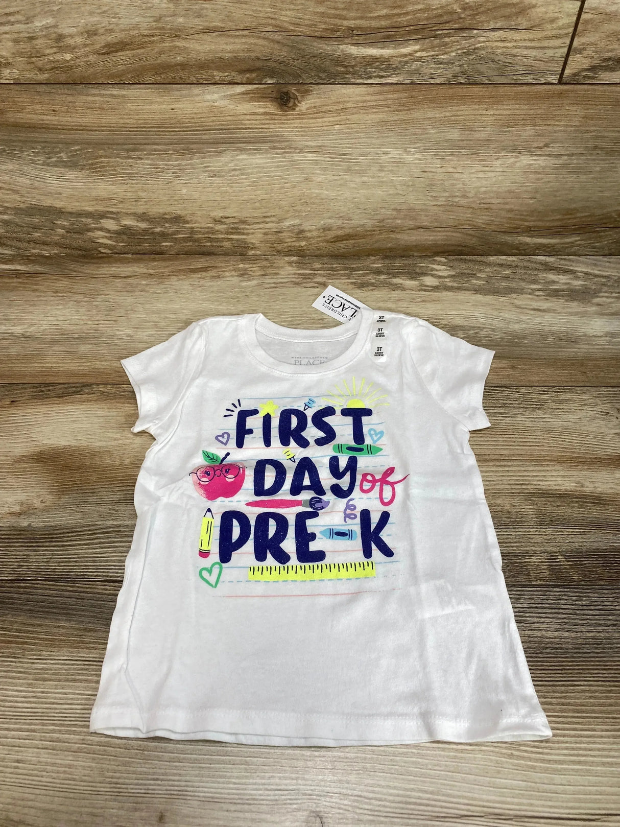 Flat lay of white Children's Place 3T short-sleeve shirt with FIRST DAY of PRE-K text and colorful school graphics on wood