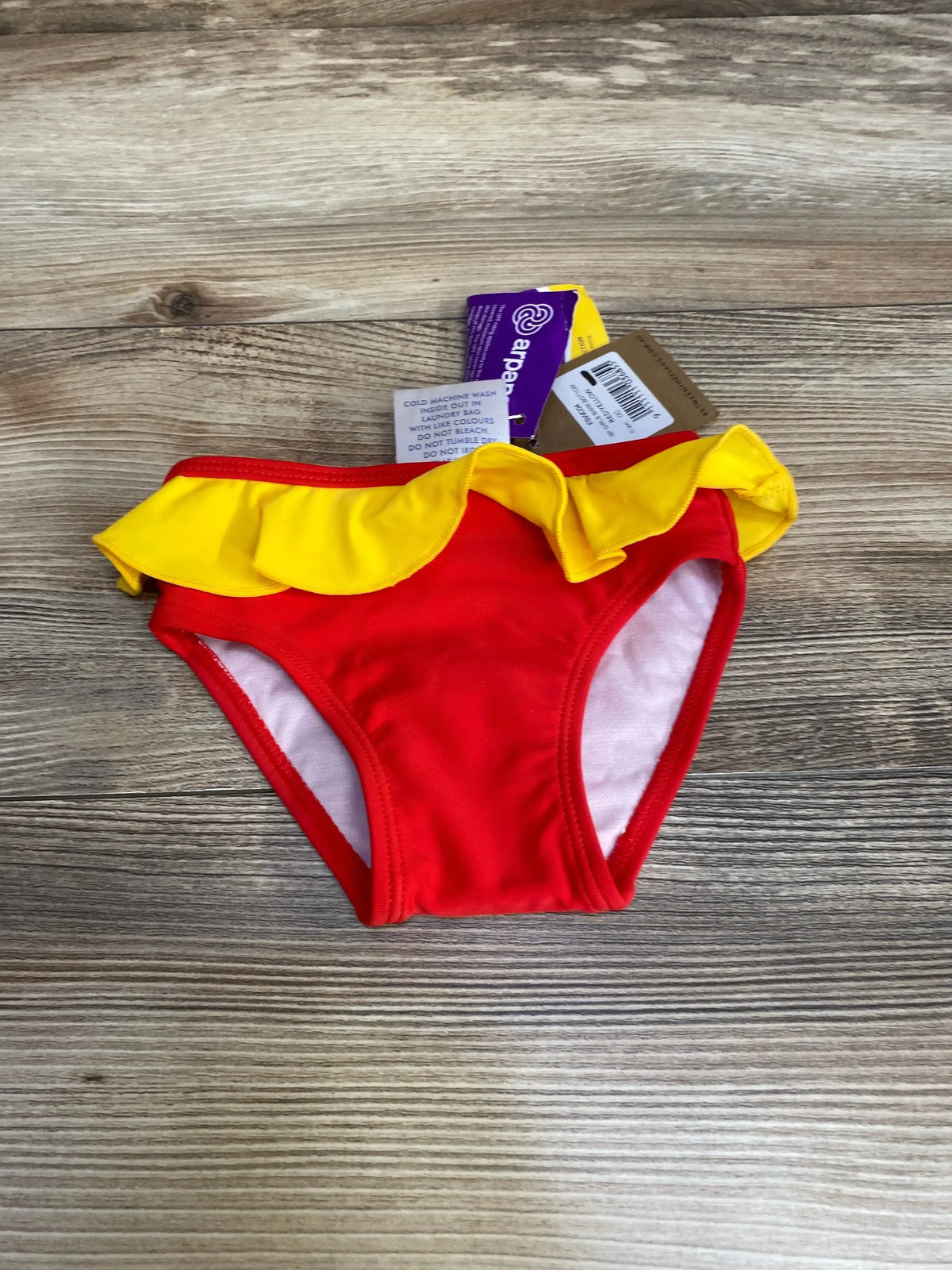Flat lay of toddler swim bottoms (size 2T) in red with a yellow ruffle waistband and visible tags on a wood background