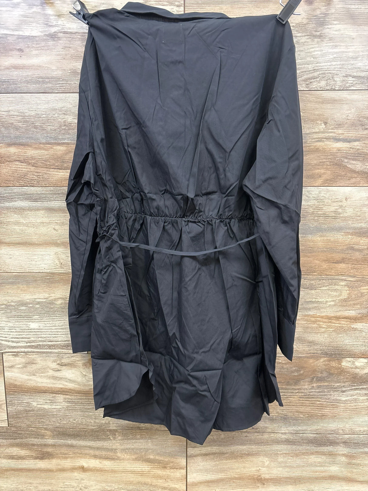 Front view of Gap maternity popover shirt in black showing long sleeves, collar, and cinched elastic waist with tie, hanging on wooden wall background
