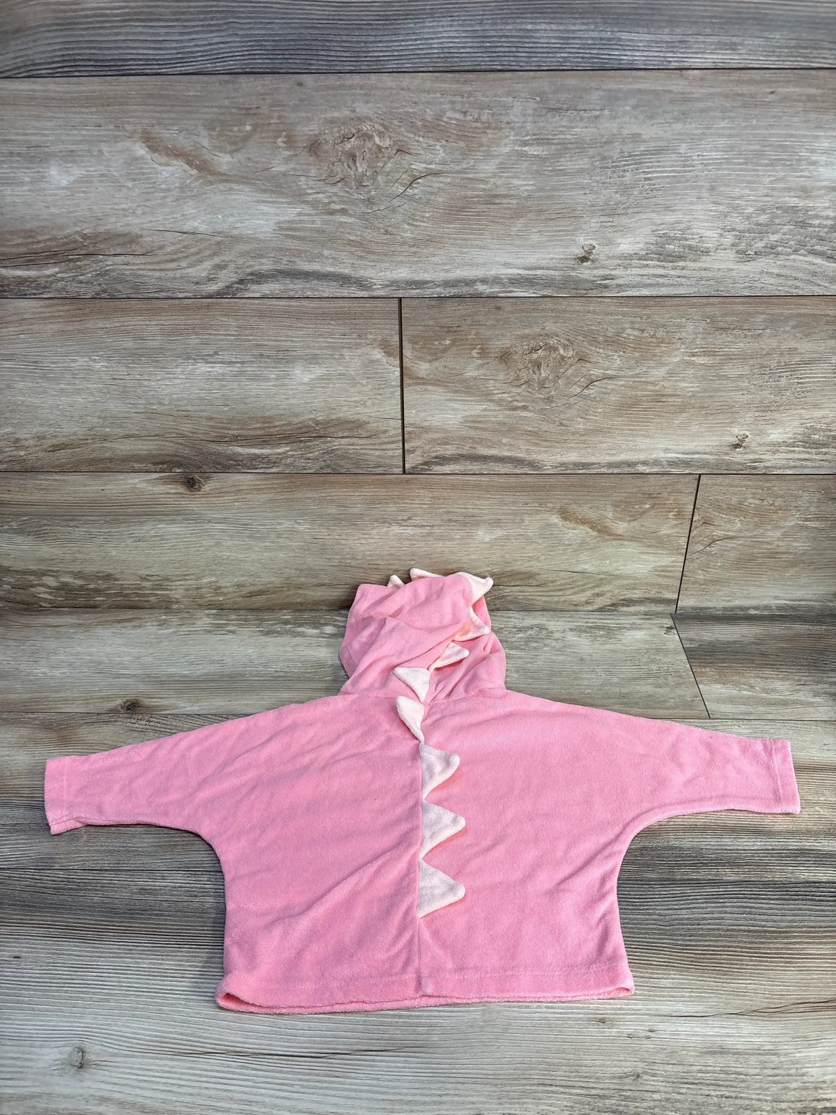 Flat lay of pink toddler swim cover-up hoodie with hood and white dinosaur spikes, wide sleeves on wood floor background