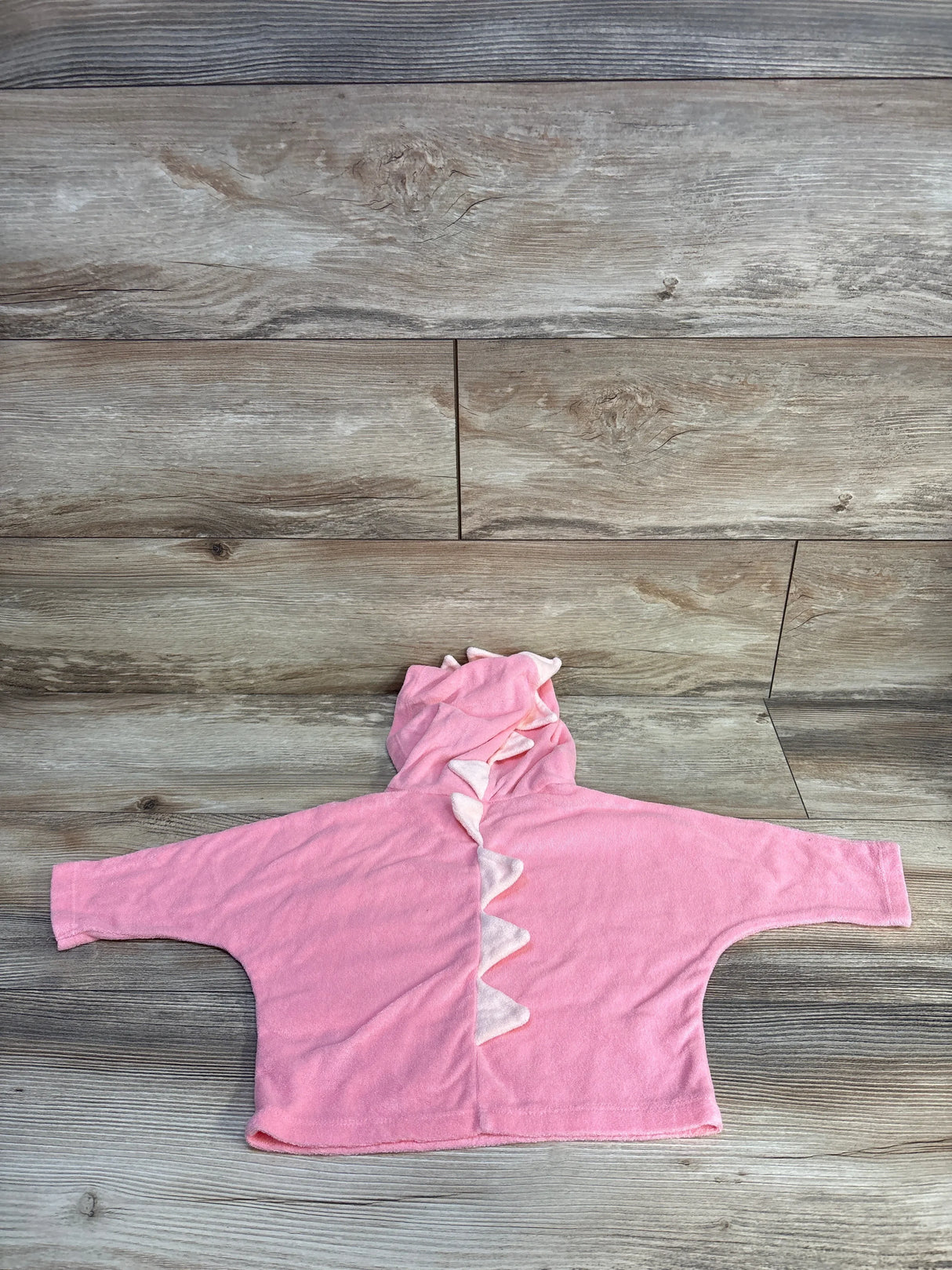 Flat lay of pink toddler swim cover-up hoodie with hood and white dinosaur spikes, wide sleeves on wood floor background