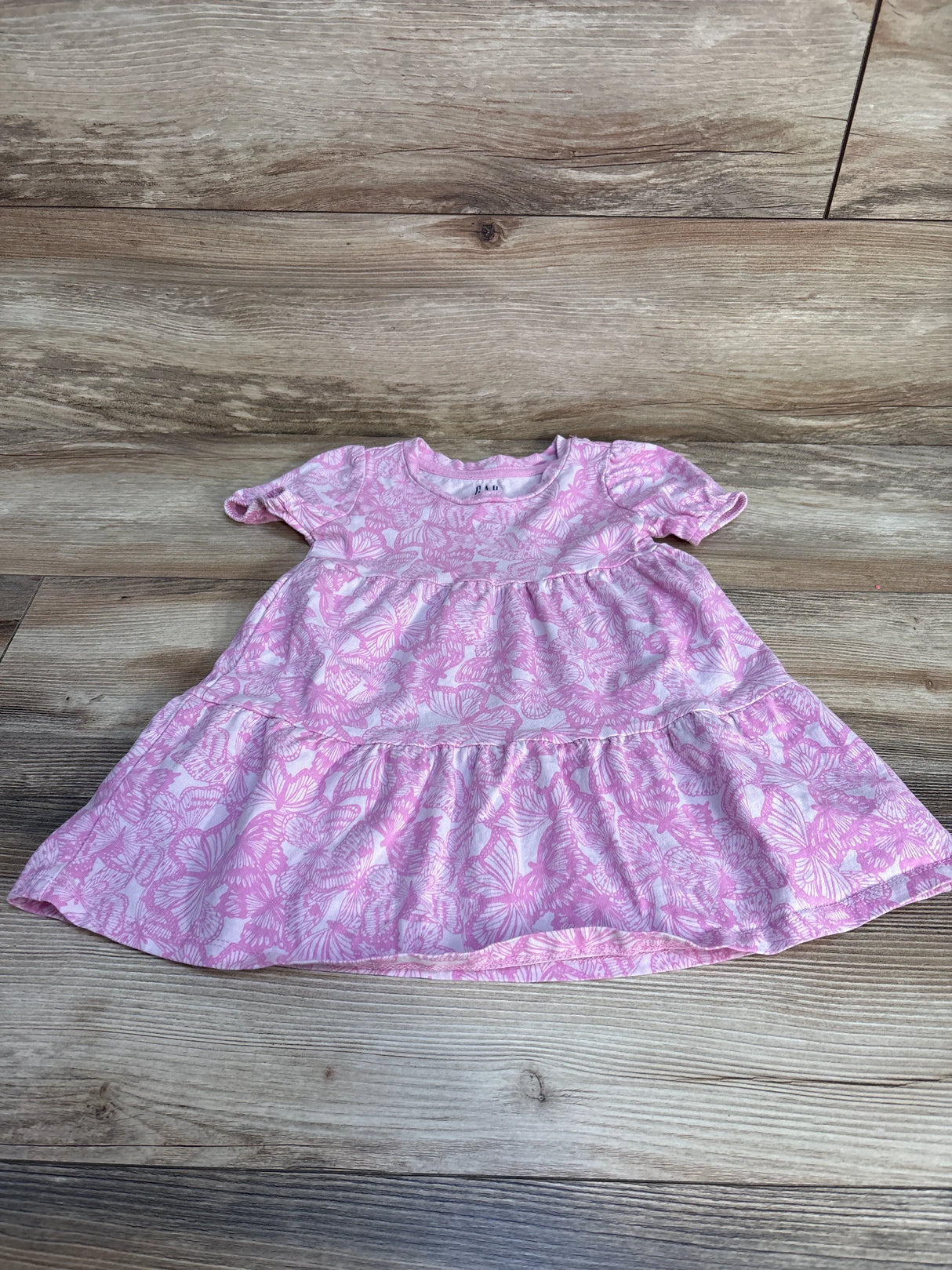 Flat lay of pink Gap Kids toddler dress with white butterfly print, short ruffle sleeves and tiered skirt on wood floor