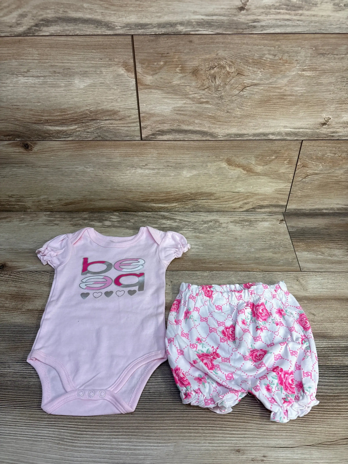 Flat lay of pink 2-piece bodysuit and floral bloomers size 6-9 months showing bebe graphic and puff sleeves on wood floor