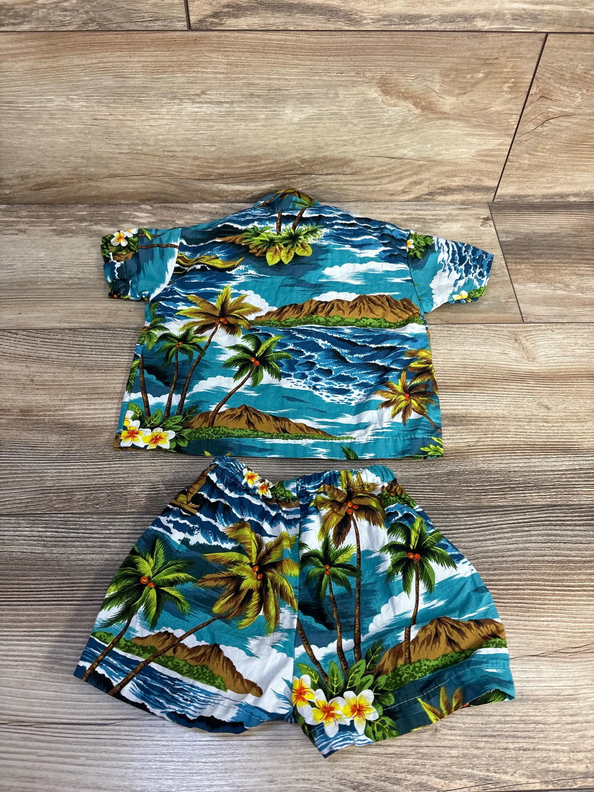 Flat lay of RJC blue two-piece button-up shirt and shorts set showcasing tropical island print on wood flooring