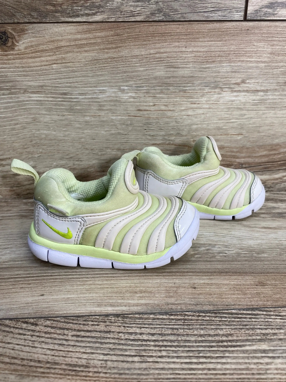 Side view of toddler Nike Dynamo Free sneakers, white and light green, showing ridged slip-on upper, pull tab, white sole on wood