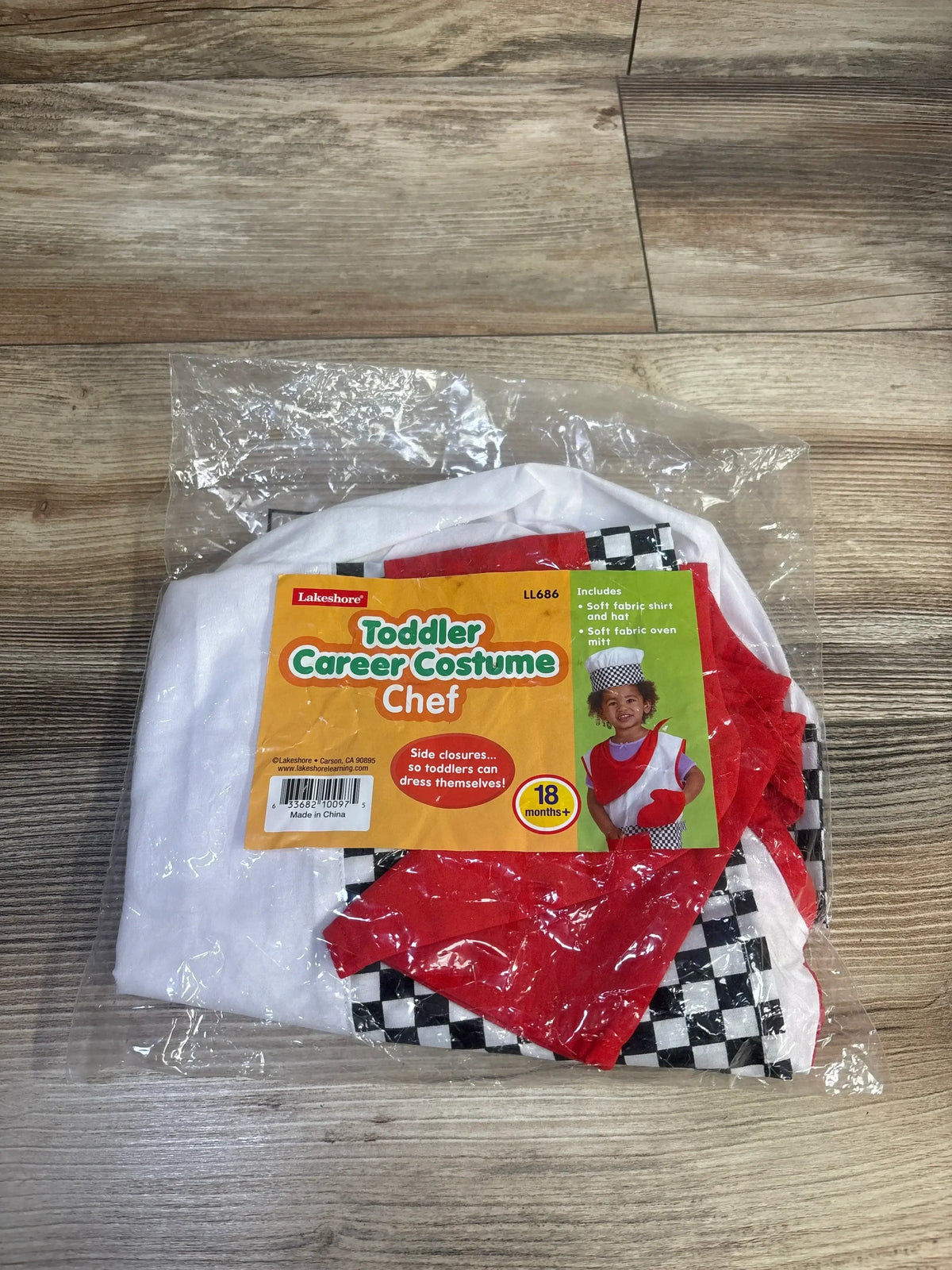 Top-down of toddler 3-piece chef costume in clear packaging: white hat, red apron, and checkered pants on wood floor